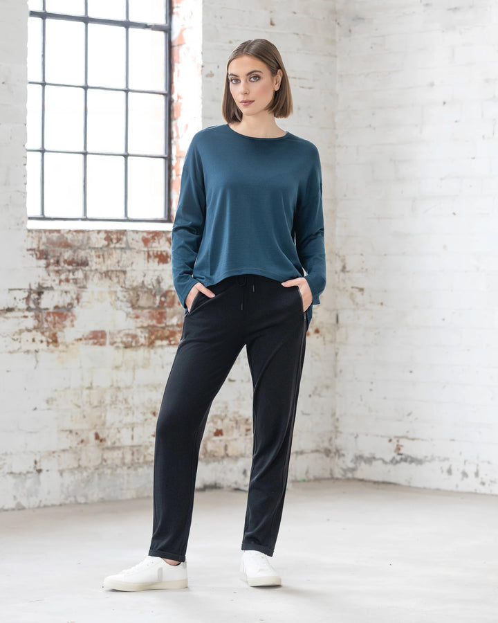 A woman stands indoors near a large window, wearing the Vassalli Quinn Merino Top in Dragonfly, paired with black pants and white sneakers. The room features white brick walls with exposed patches and a concrete floor.