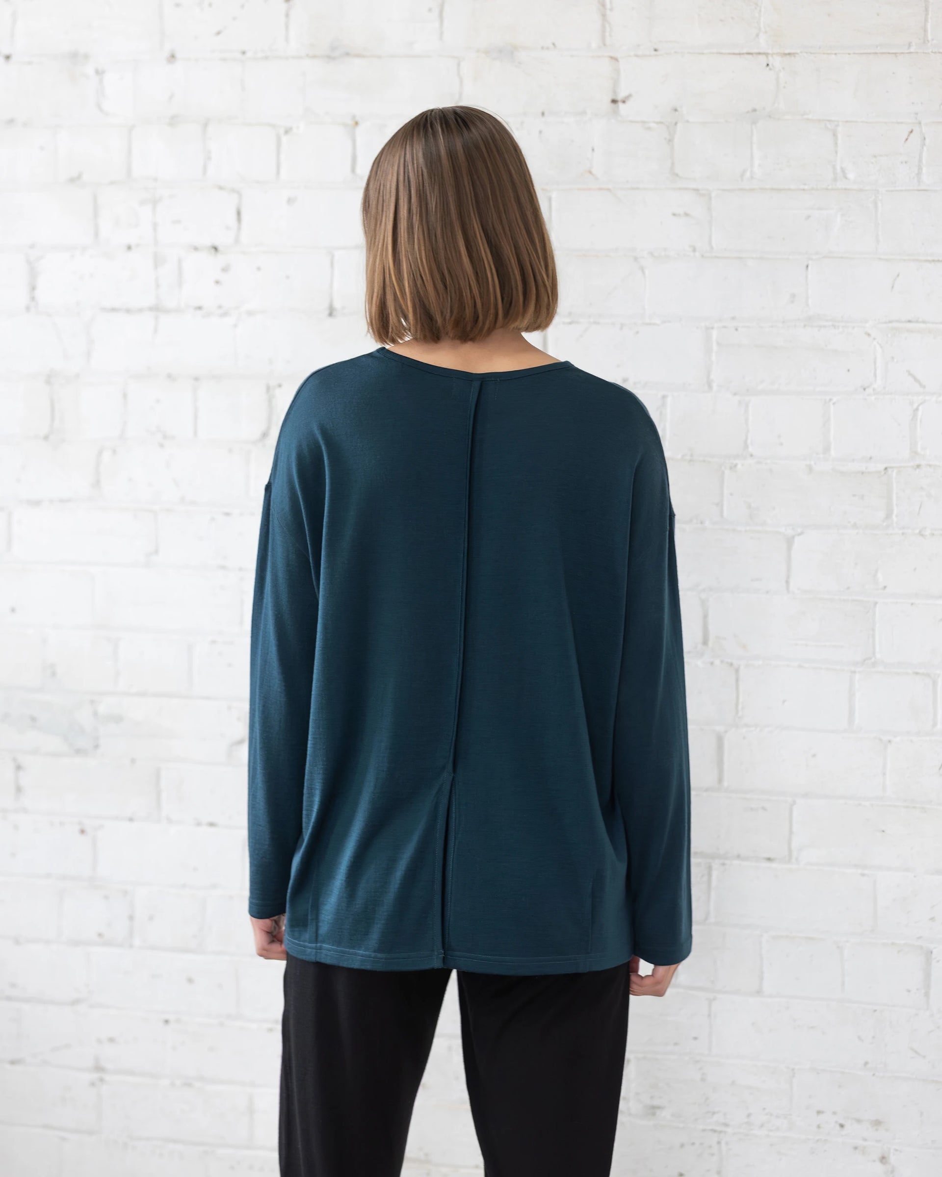 A person with straight, shoulder-length brown hair faces a white brick wall, wearing the Vassalli Quinn Merino Top in Dragonfly—an oversized merino piece with visible back seam detail—paired with black pants.