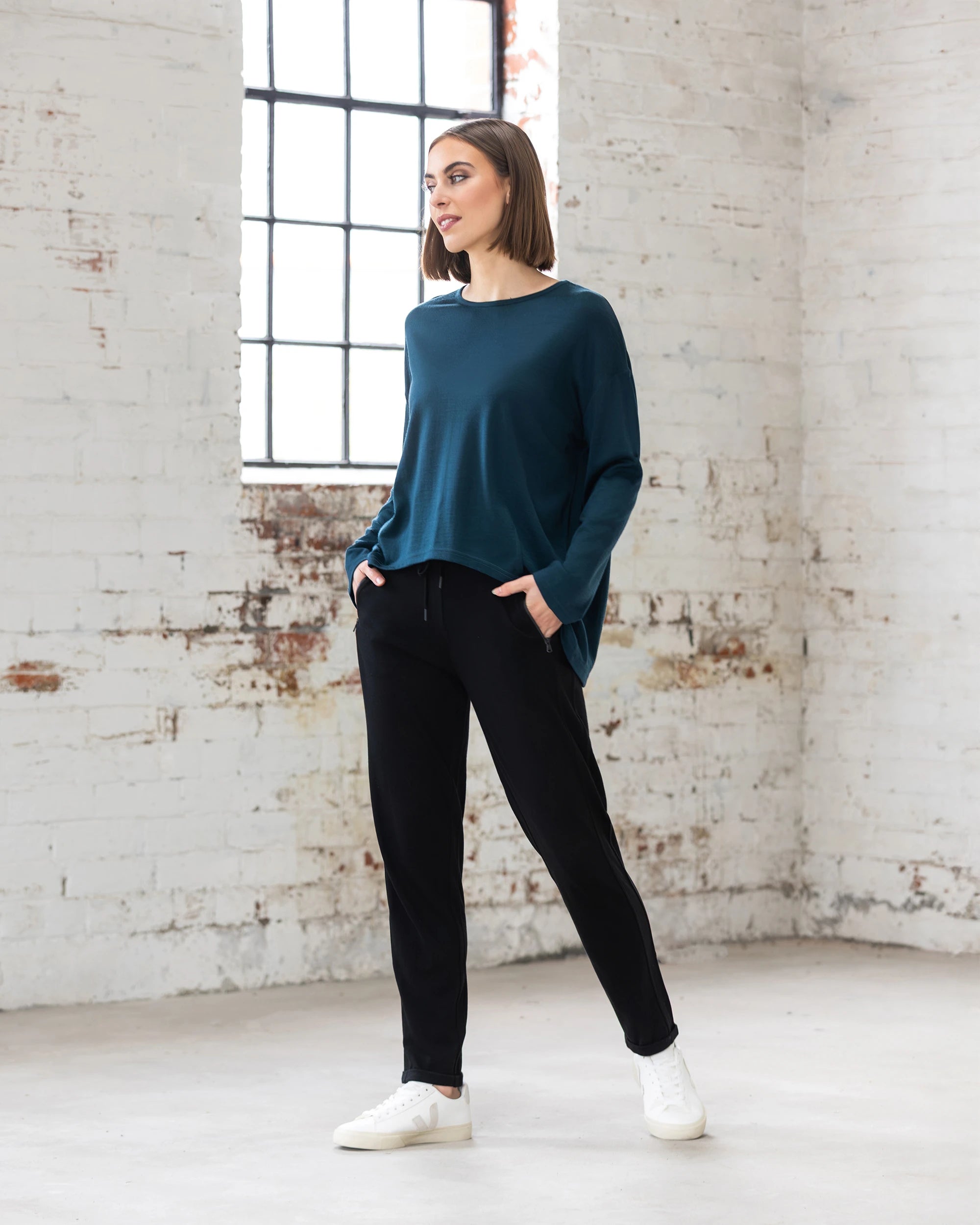 A woman stands indoors near a large window, wearing the Vassalli Quinn Merino Top in Dragonfly with black pants and white sneakers. Hands in pockets, she smiles slightly, showcasing modern merino style against a white brick wall.