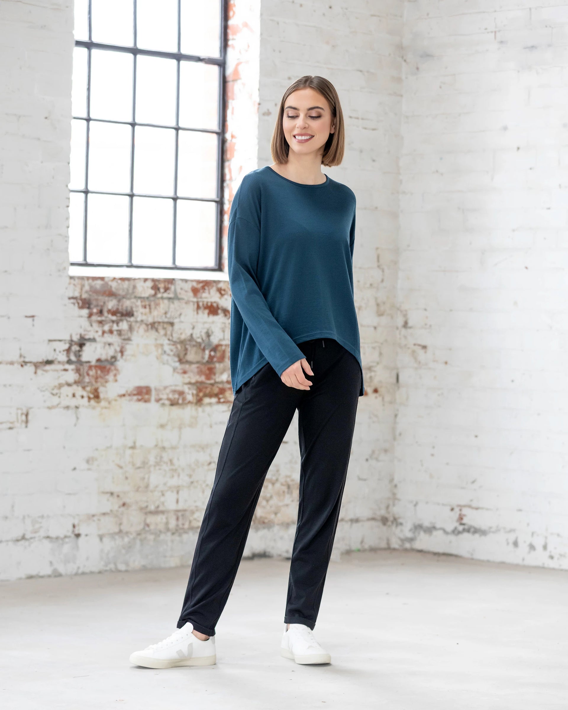 A woman smiles to the side in a bright, minimalist room wearing the Vassalli Quinn Merino Top in Dragonfly, paired with black pants and white sneakers—exemplifying effortless modern merino style.