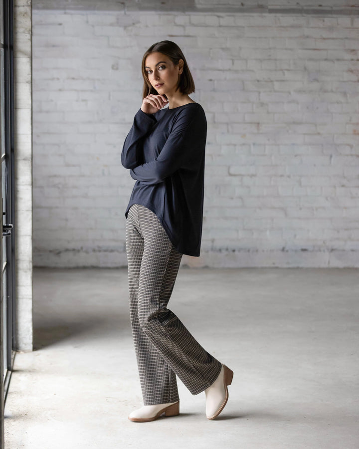 A woman stands in a minimalist room with white brick walls, thoughtfully posing in the Vassalli Quinn Merino Top - Midnight, paired with checked pants and beige shoes.