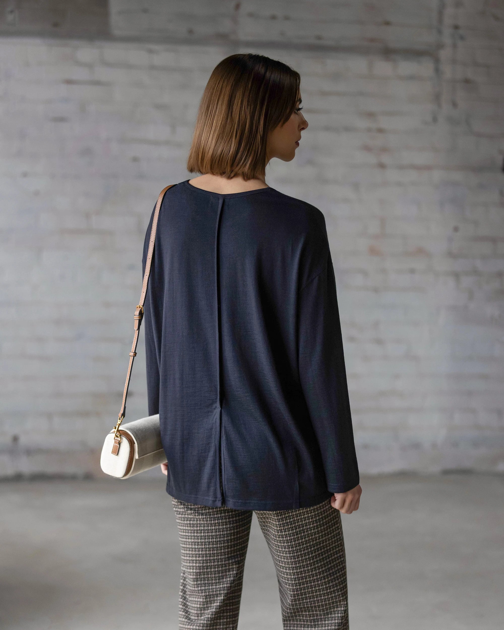 A woman seen from behind wears the Vassalli Quinn Merino Top in Midnight, styled with grey plaid trousers and a small cream crossbody bag featuring a tan leather strap.