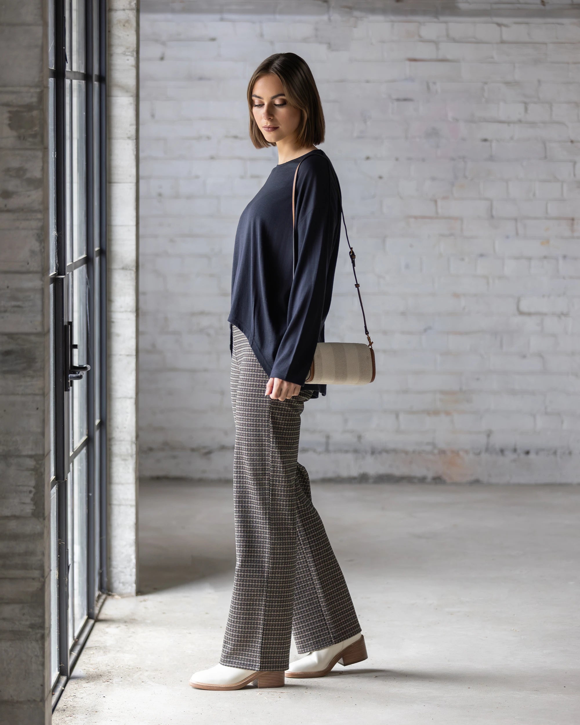 A woman with short hair and a neutral expression stands indoors by a large window, wearing the Vassalli Quinn Merino Top in Midnight, plaid pants, cream shoes, and a small shoulder bag against a white brick wall.