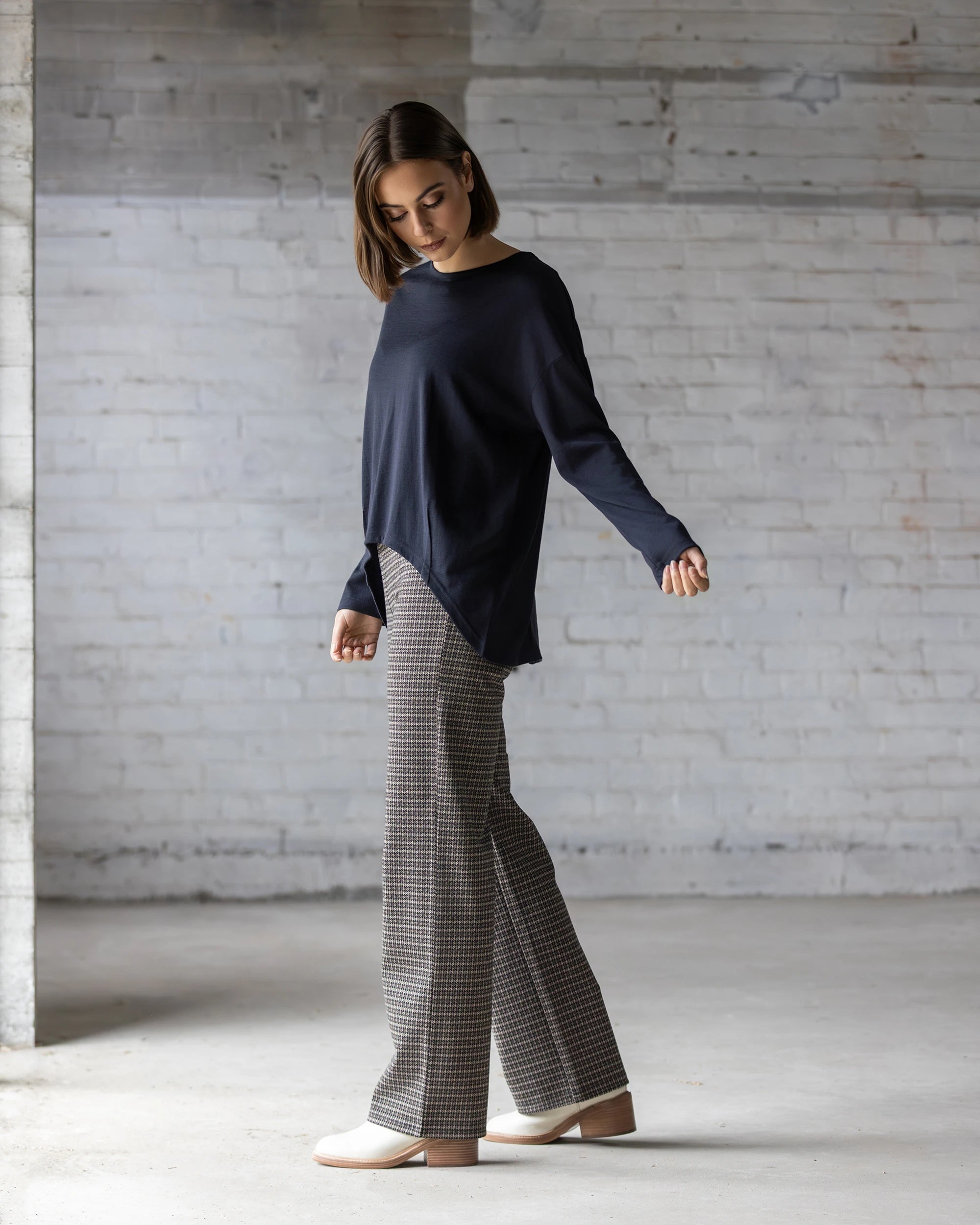 A person with short brown hair wears the Vassalli Quinn Merino Top in Midnight, plaid wide-leg pants, and beige shoes, standing in a minimal, light-filled room with white brick walls and looking down.