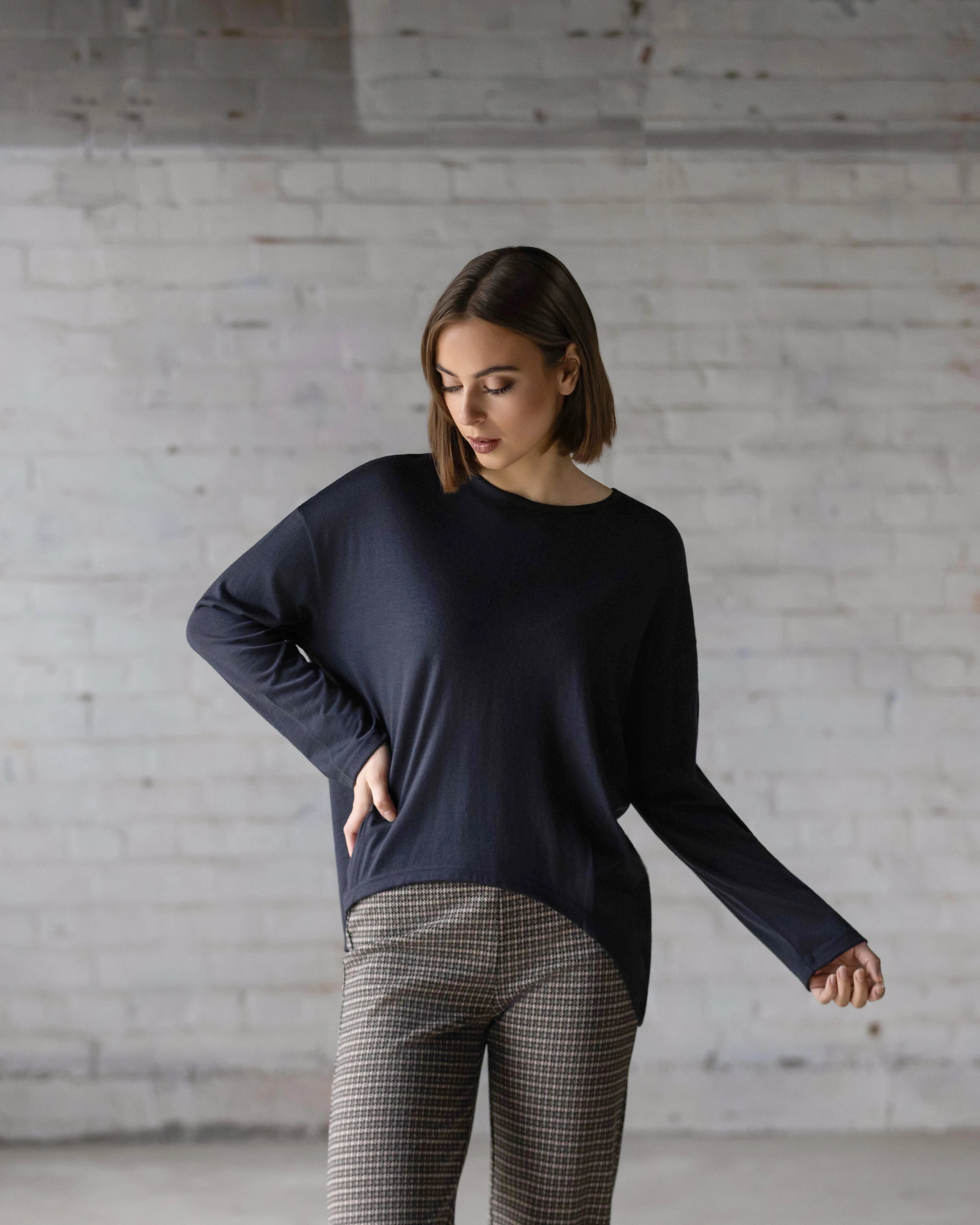 A woman with short brown hair stands against a white brick wall, looking down. She wears the oversized Quinn Merino Top - Midnight by Vassalli and patterned pants, with one arm bent and the other hanging by her side.