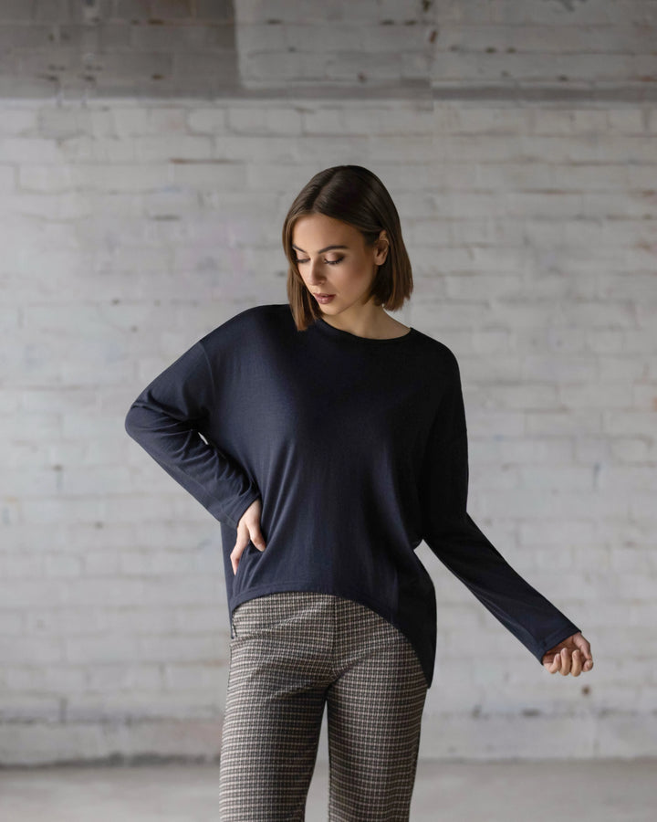 A woman with short brown hair stands against a white brick wall, looking down. She wears the oversized Quinn Merino Top - Midnight by Vassalli and patterned pants, with one arm bent and the other hanging by her side.