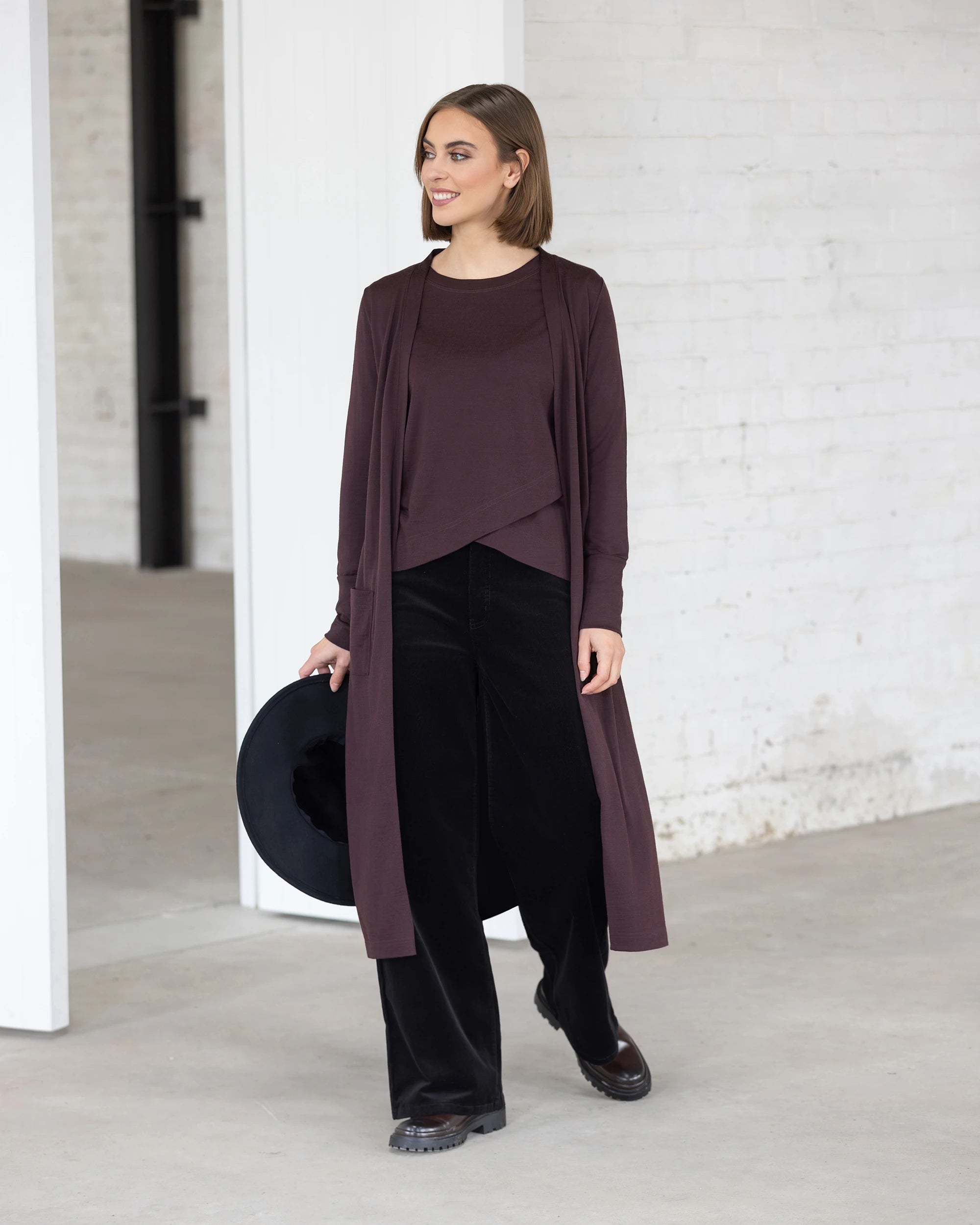 A woman stands indoors by white brick walls and a large doorway, smiling as she models the Vassalli Suki Merino Coatigan in Berry over a matching top and black wide-leg pants, holding a black hat in one hand.