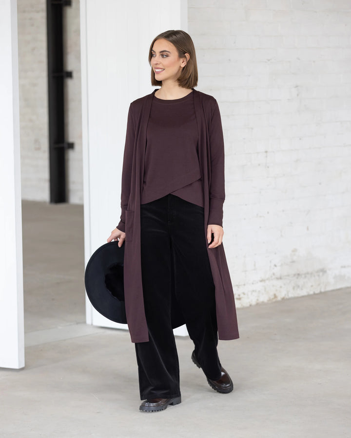 A woman stands indoors by white brick walls and a large doorway, smiling as she models the Vassalli Suki Merino Coatigan in Berry over a matching top and black wide-leg pants, holding a black hat in one hand.