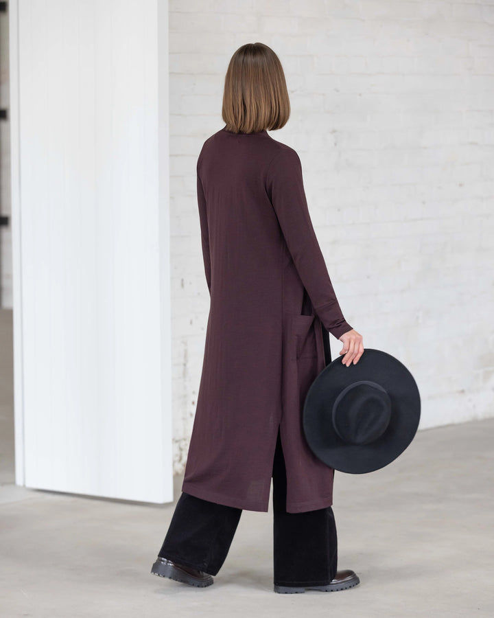 A person with straight, shoulder-length hair stands indoors with their back to the camera, wearing the Vassalli Suki Merino Coatigan in Berry over black pants and holding a wide-brimmed black hat by their side.