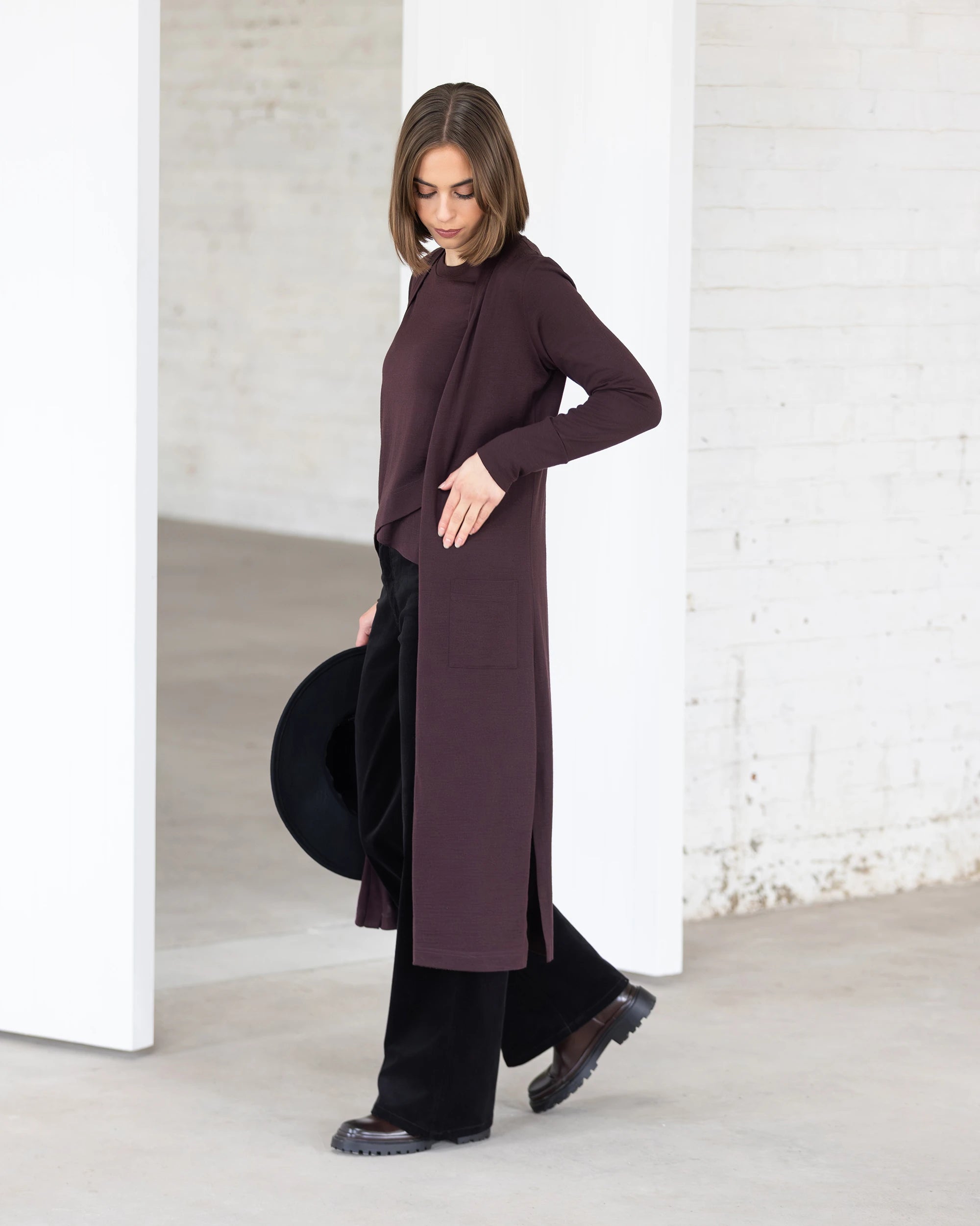 A woman stands indoors looking down, holding a black hat and wearing the Vassalli Suki Merino Coatigan in Berry with black pants and shoes. The bright room with white walls highlights this cozy layering piece perfectly.
