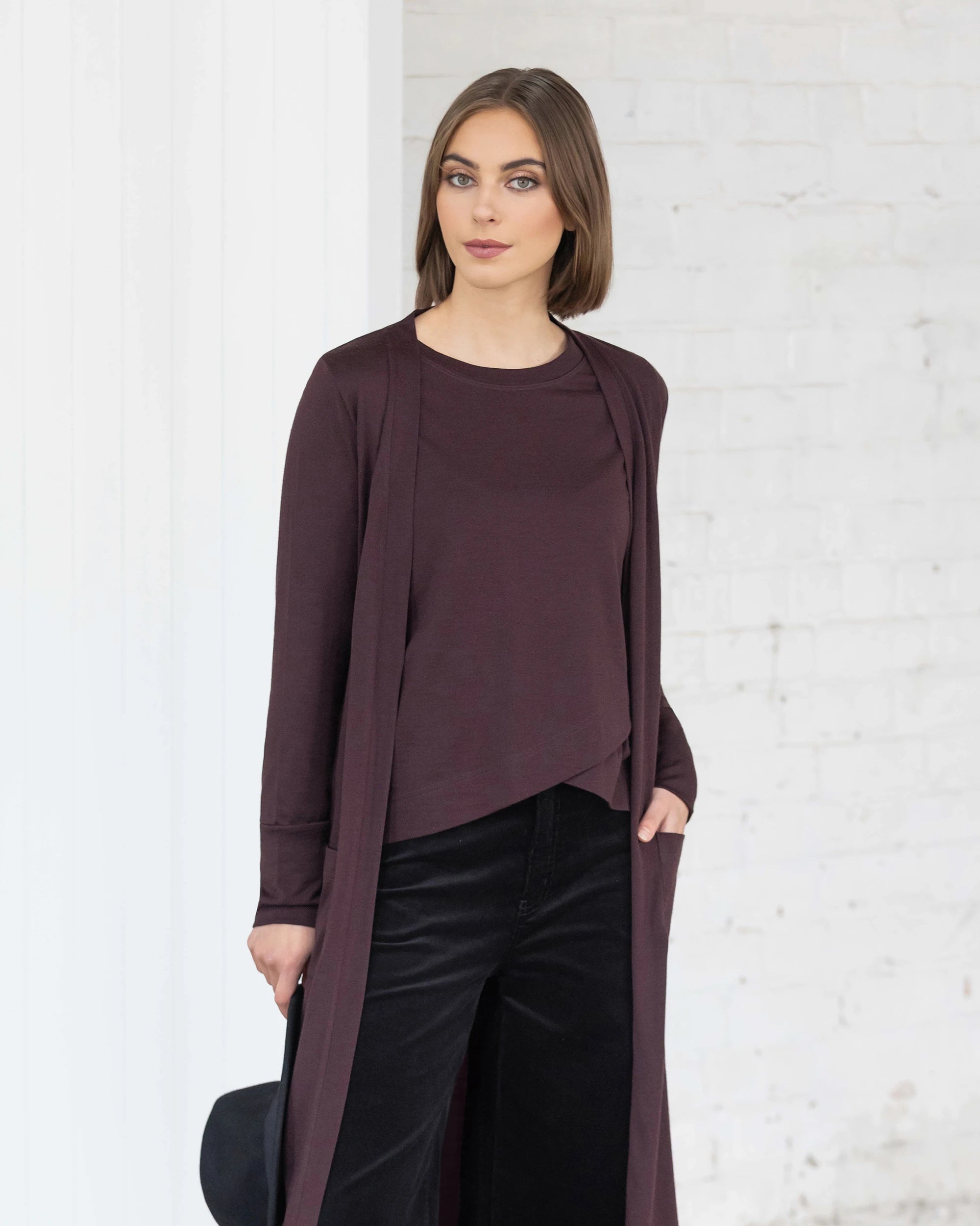 A woman with straight brown hair wears the Vassalli Suki Merino Coatigan in berry over a matching top and black pants, standing against a white brick wall and holding a black hat while looking at the camera.