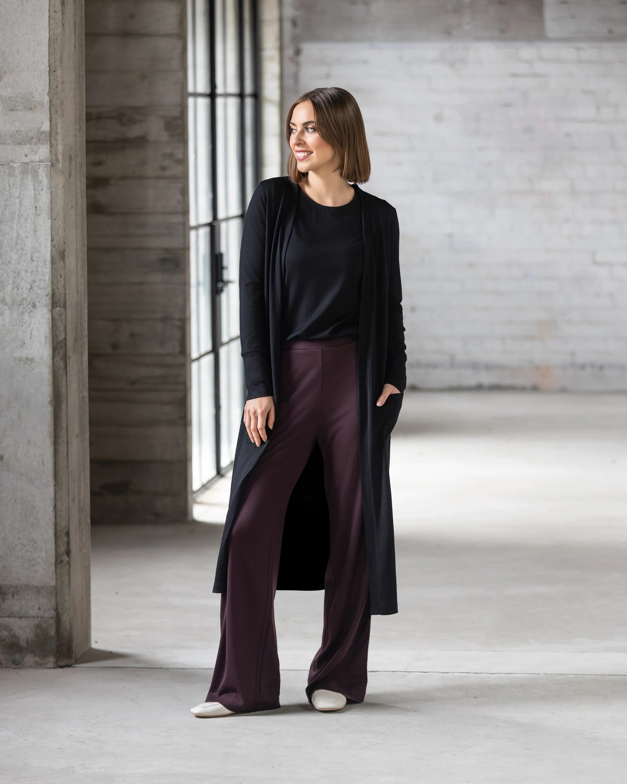 A woman with chin-length brown hair stands indoors by large windows, smiling to the side. She wears the Vassalli Suki Merino Coatigan in black over a black top, maroon wide-leg pants, and white shoes.