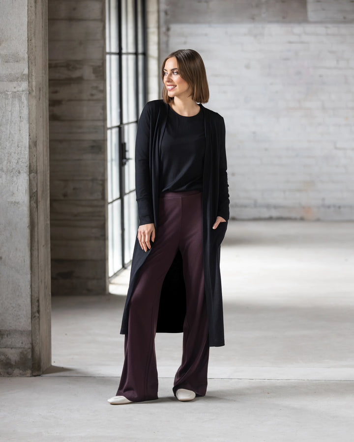 A woman with chin-length brown hair stands indoors by large windows, smiling to the side. She wears the Vassalli Suki Merino Coatigan in black over a black top, maroon wide-leg pants, and white shoes.