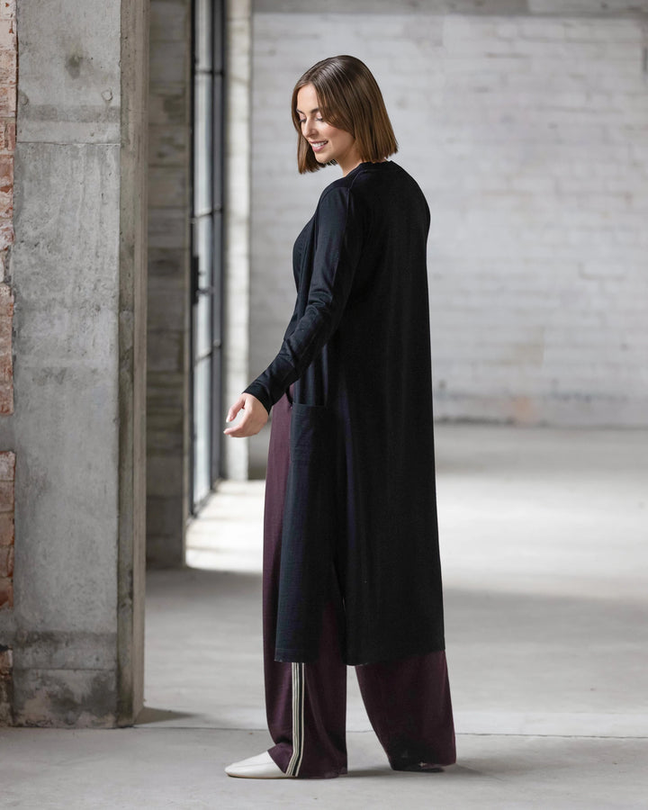 A woman with chin-length brown hair, wearing the Vassalli Suki Merino Coatigan in black, loose maroon pants, and white shoes stands indoors in a bright, industrial-style space, looking back over her shoulder.
