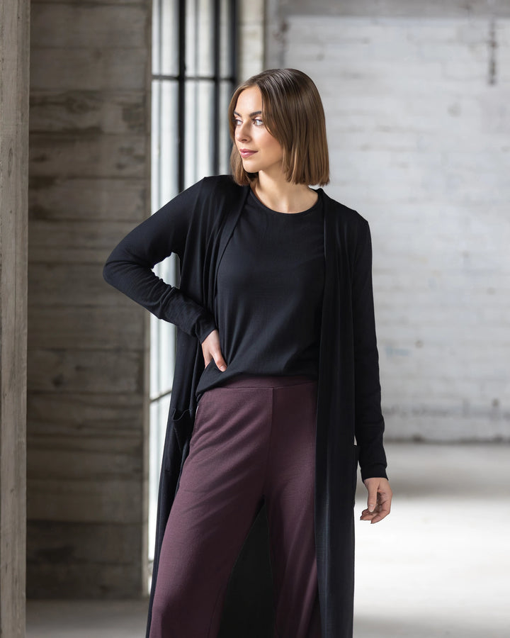 A woman with shoulder-length brown hair wears the Vassalli Suki Merino Coatigan in black, paired with a black top and burgundy pants, standing indoors by large windows—a chic and versatile layering essential.