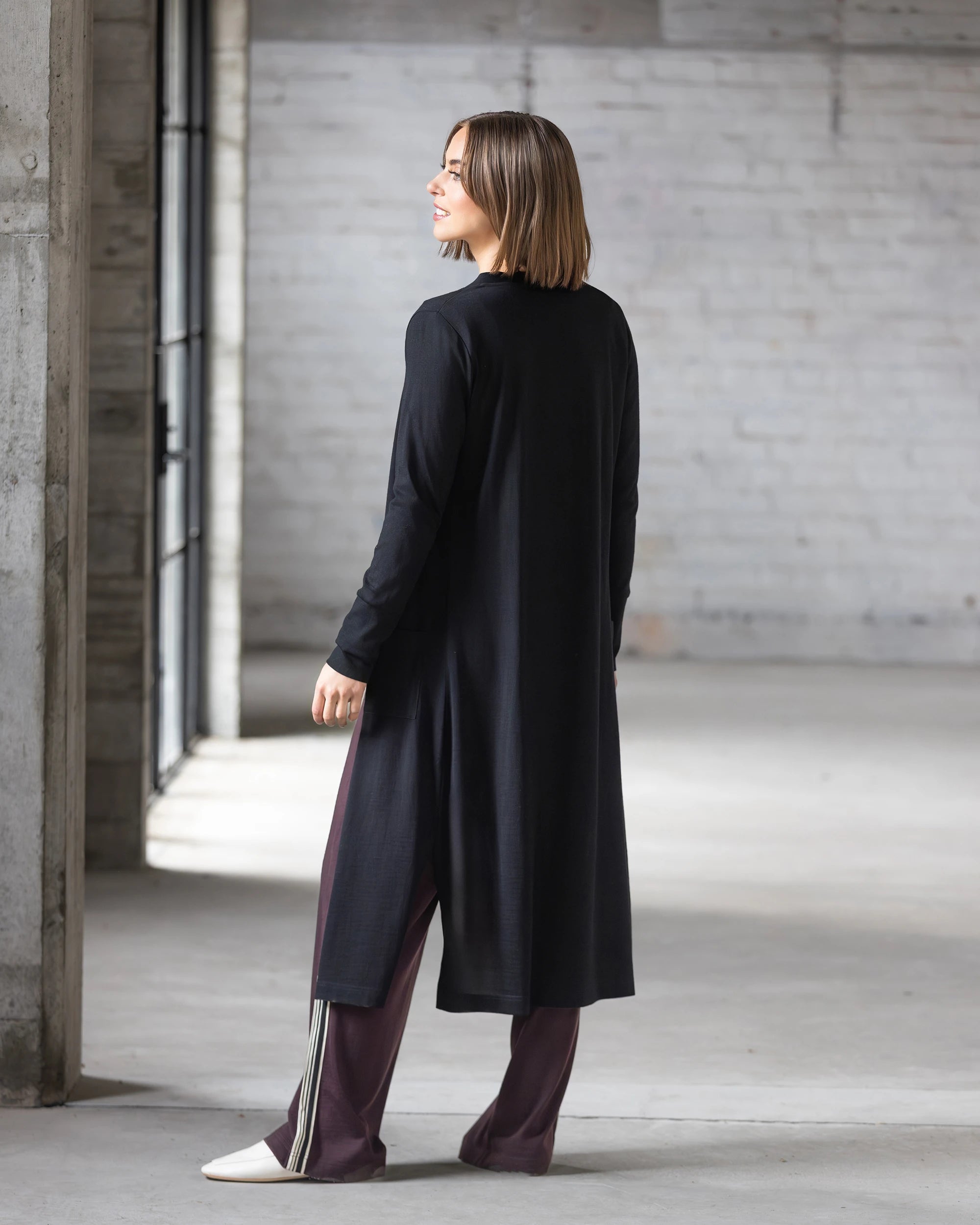 A woman with shoulder-length brown hair stands in a minimalist, industrial-style room, wearing the Vassalli Suki Merino Coatigan in black over wide-legged purple pants and white shoes. She faces away while looking to the side.