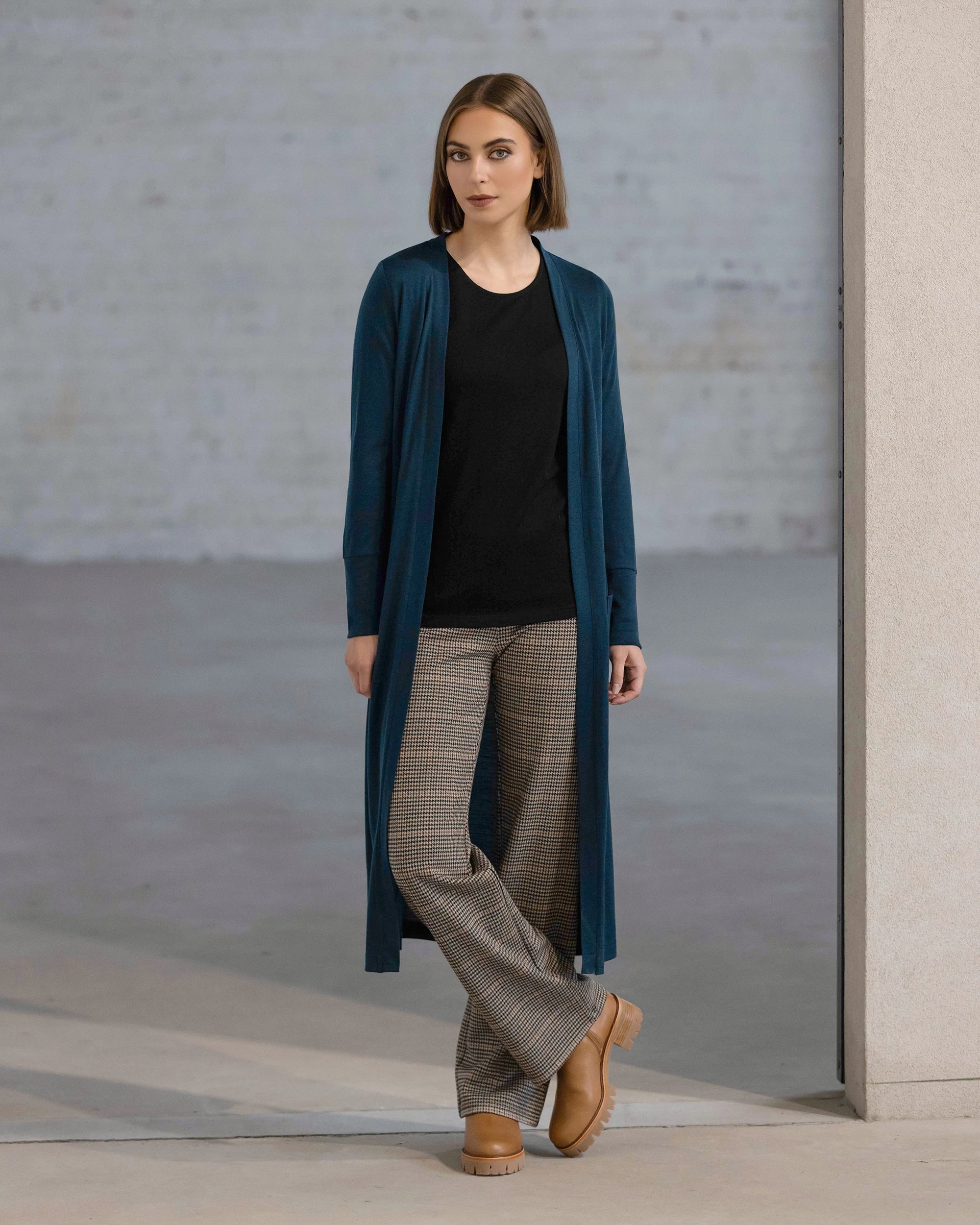 A woman wears the Vassalli Suki Merino Coatigan – Dragonfly over a black top, plaid trousers, and tan platform shoes, standing indoors against a light wall and gray background with a neutral expression.