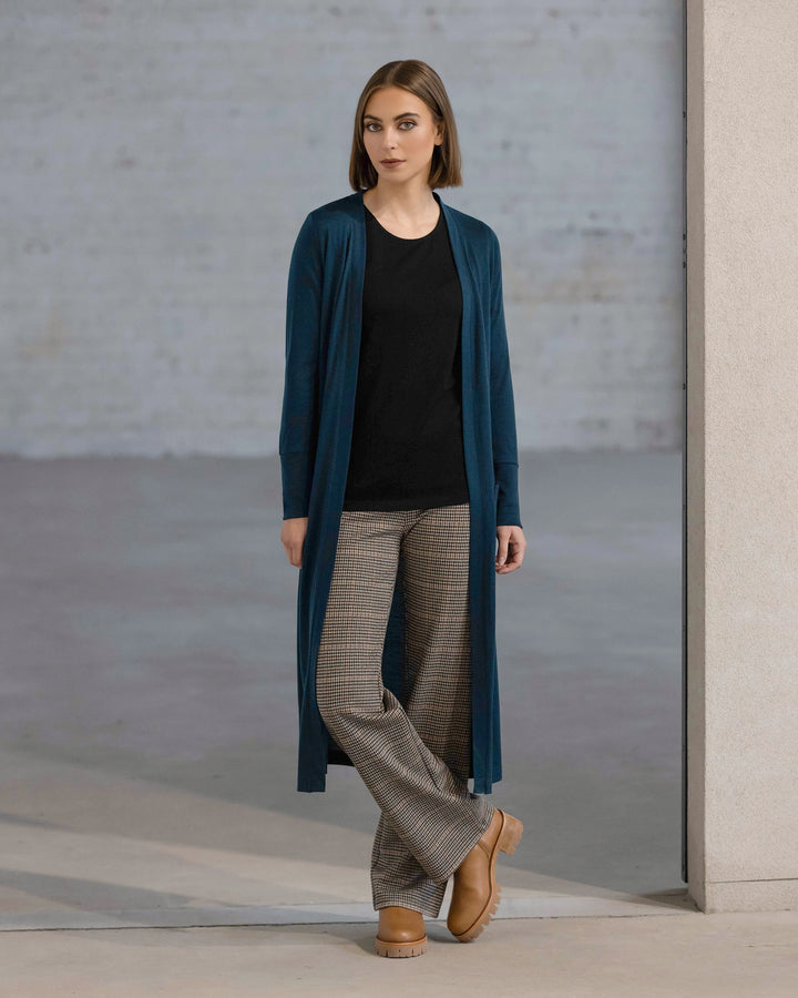 A woman wears the Vassalli Suki Merino Coatigan – Dragonfly over a black top, plaid trousers, and tan platform shoes, standing indoors against a light wall and gray background with a neutral expression.