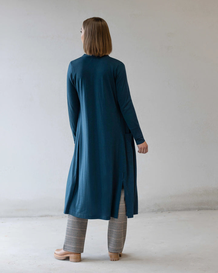 A person stands indoors facing away, wearing the Vassalli Suki Merino Coatigan in Dragonfly over gray checkered pants and light platform shoes, with a plain light-colored wall and floor in the background.