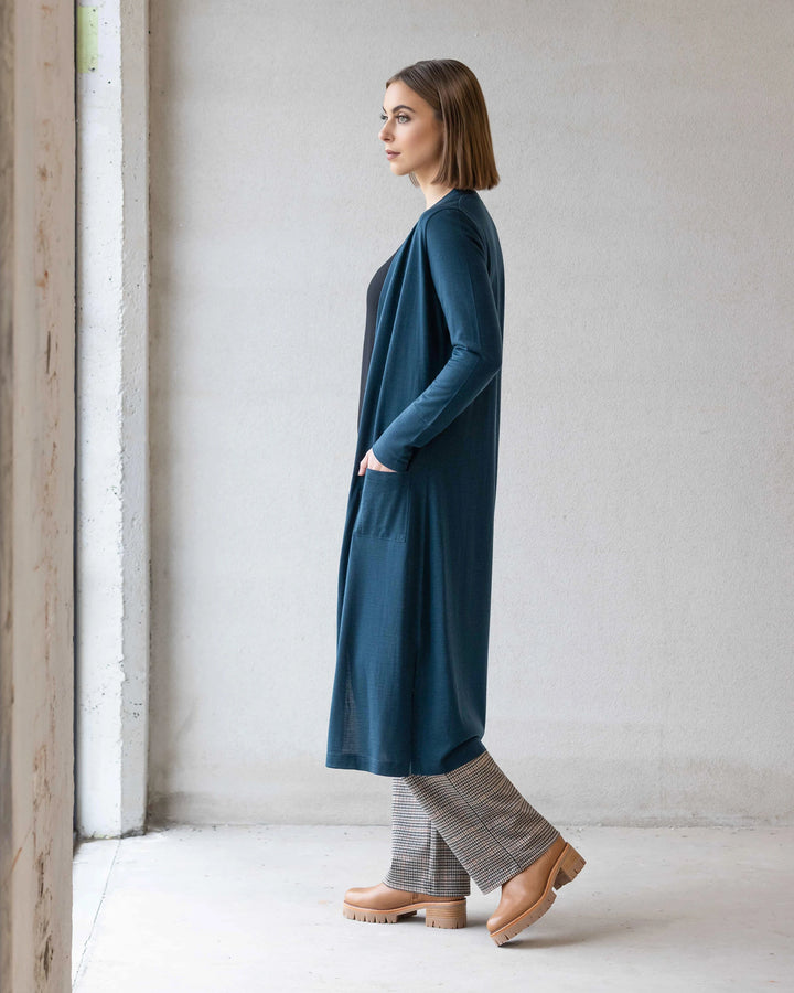 A woman with straight, shoulder-length hair stands sideways indoors, wearing the Vassalli Suki Merino Coatigan - Dragonfly over a black top and plaid pants with beige platform shoes, against a light-colored wall.