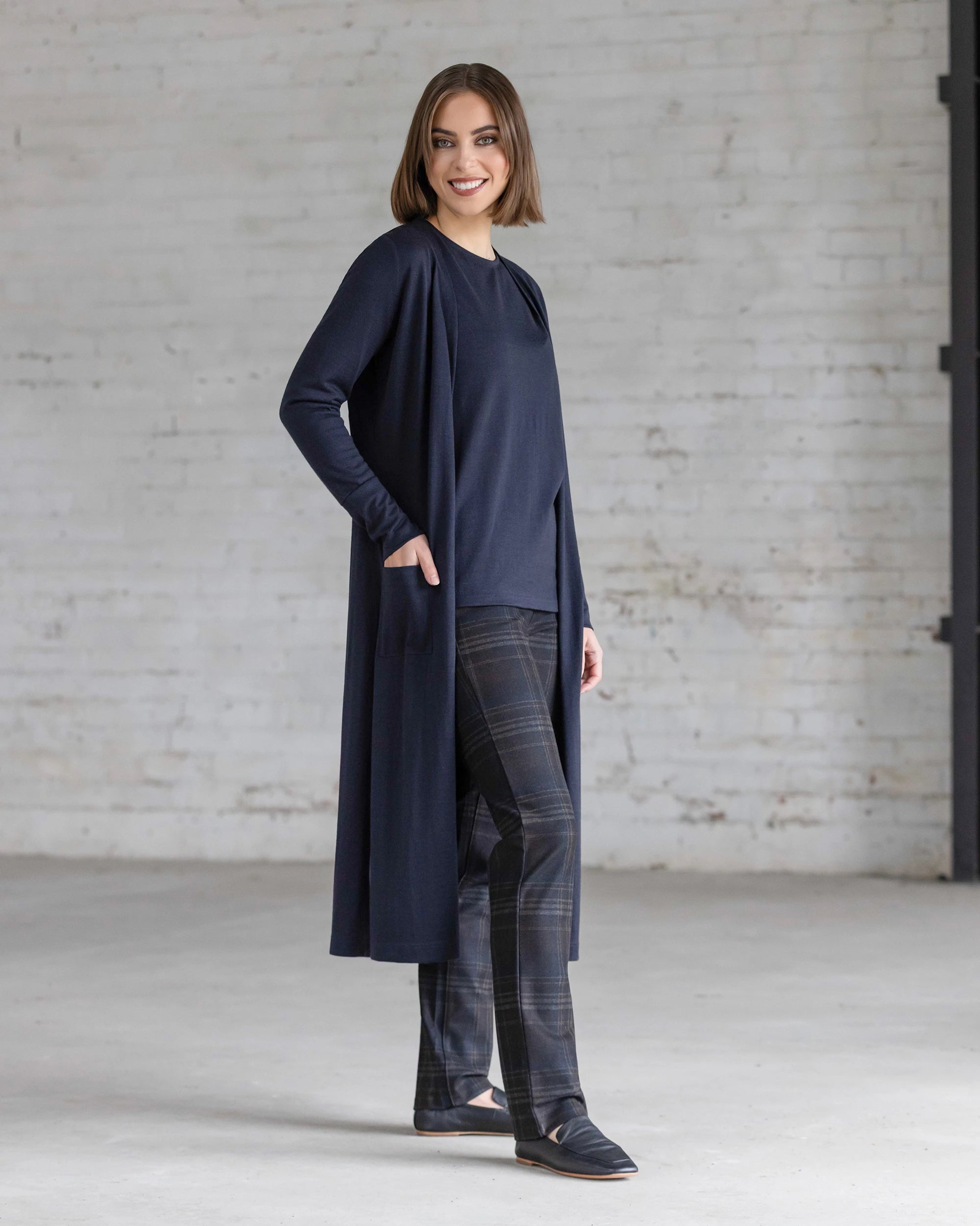 A woman stands indoors against a white brick wall, smiling as she wears the Vassalli Suki Merino Coatigan in Midnight over a matching top, paired with plaid pants and black shoes. One hand rests casually in her coatigan pocket.