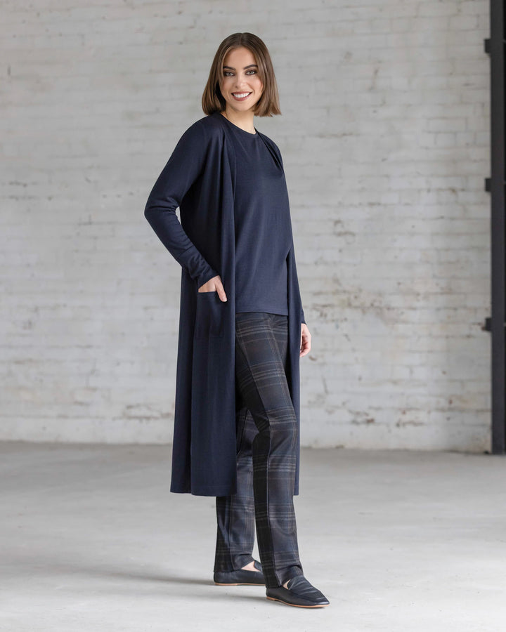 A woman stands indoors against a white brick wall, smiling as she wears the Vassalli Suki Merino Coatigan in Midnight over a matching top, paired with plaid pants and black shoes. One hand rests casually in her coatigan pocket.