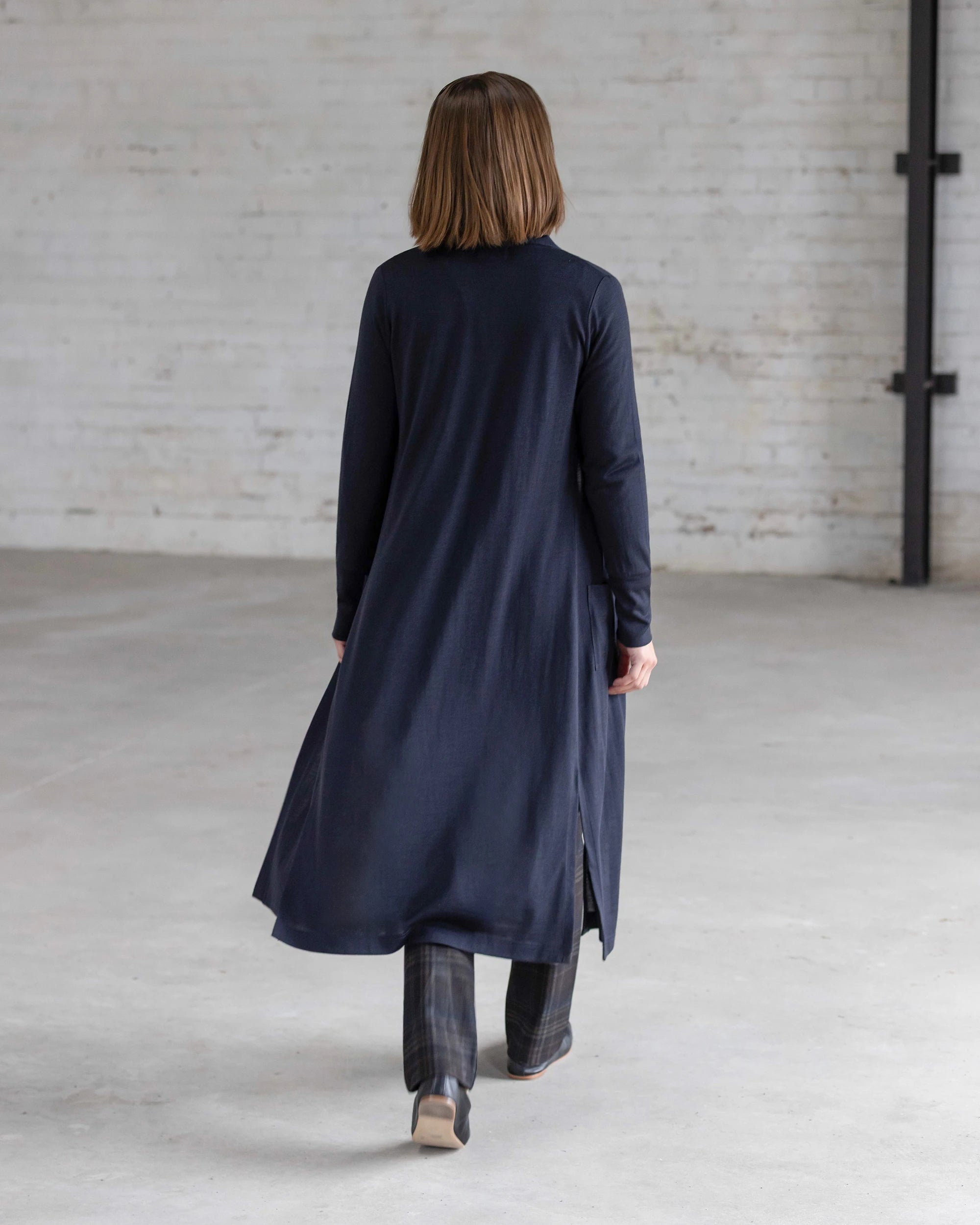 Back view of a woman in the Vassalli Suki Merino Coatigan in Midnight, featuring side slits, layered over grey plaid wide-leg trousers and dark heeled ankle boots.
