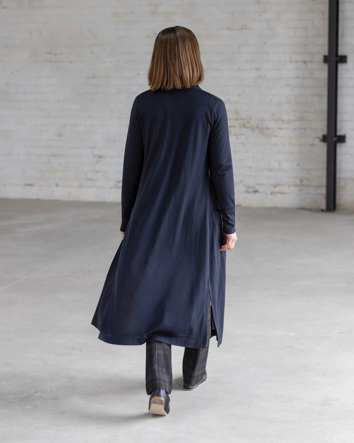 Back view of a woman in the Vassalli Suki Merino Coatigan in Midnight, featuring side slits, layered over grey plaid wide-leg trousers and dark heeled ankle boots.