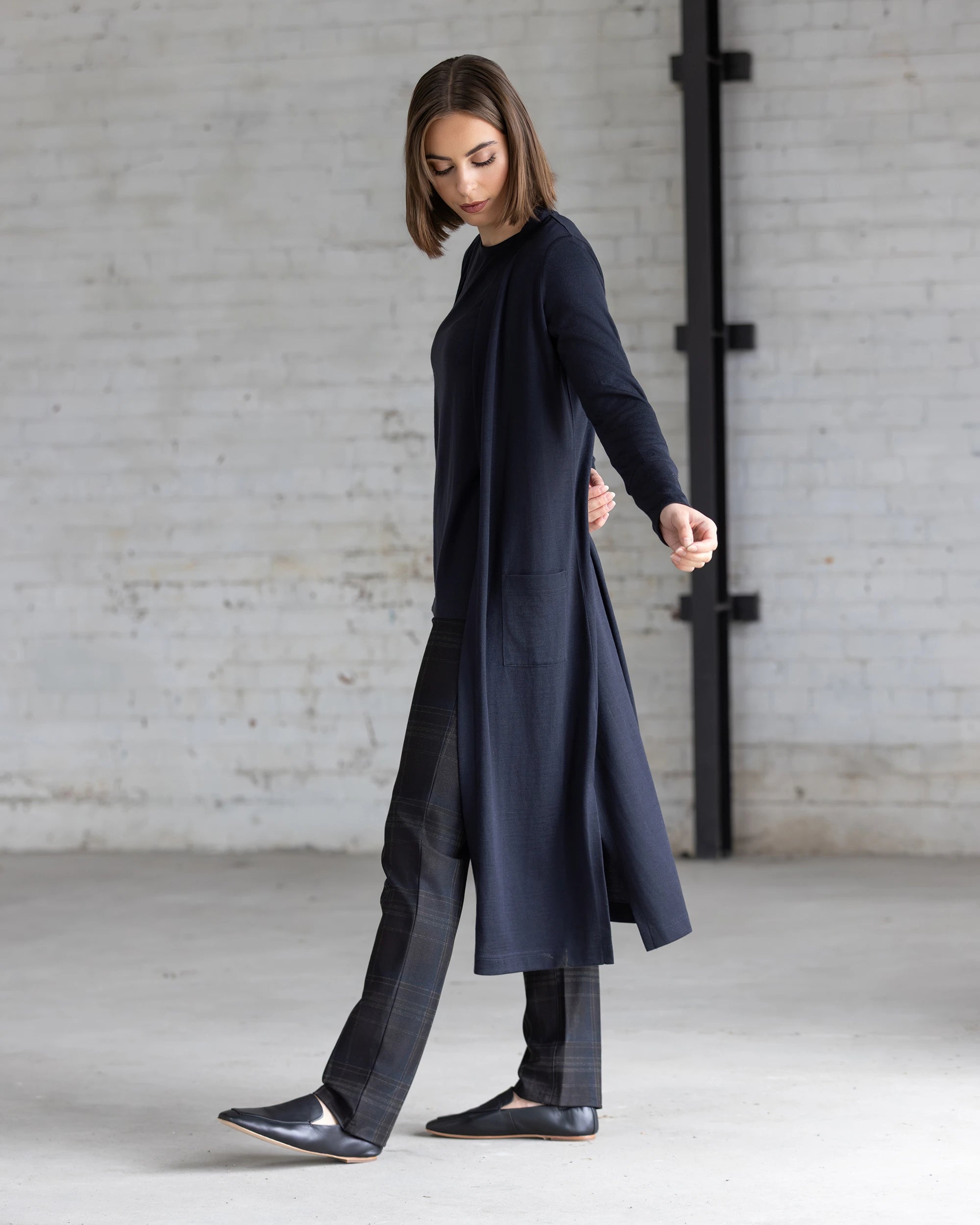 In a minimal, industrial-style room with white brick walls, a woman wears the Vassalli Suki Merino Coatigan in Midnight over plaid pants and black shoes, looking down with one arm slightly outstretched.