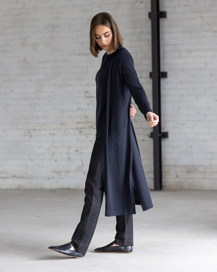 In a minimal, industrial-style room with white brick walls, a woman wears the Vassalli Suki Merino Coatigan in Midnight over plaid pants and black shoes, looking down with one arm slightly outstretched.