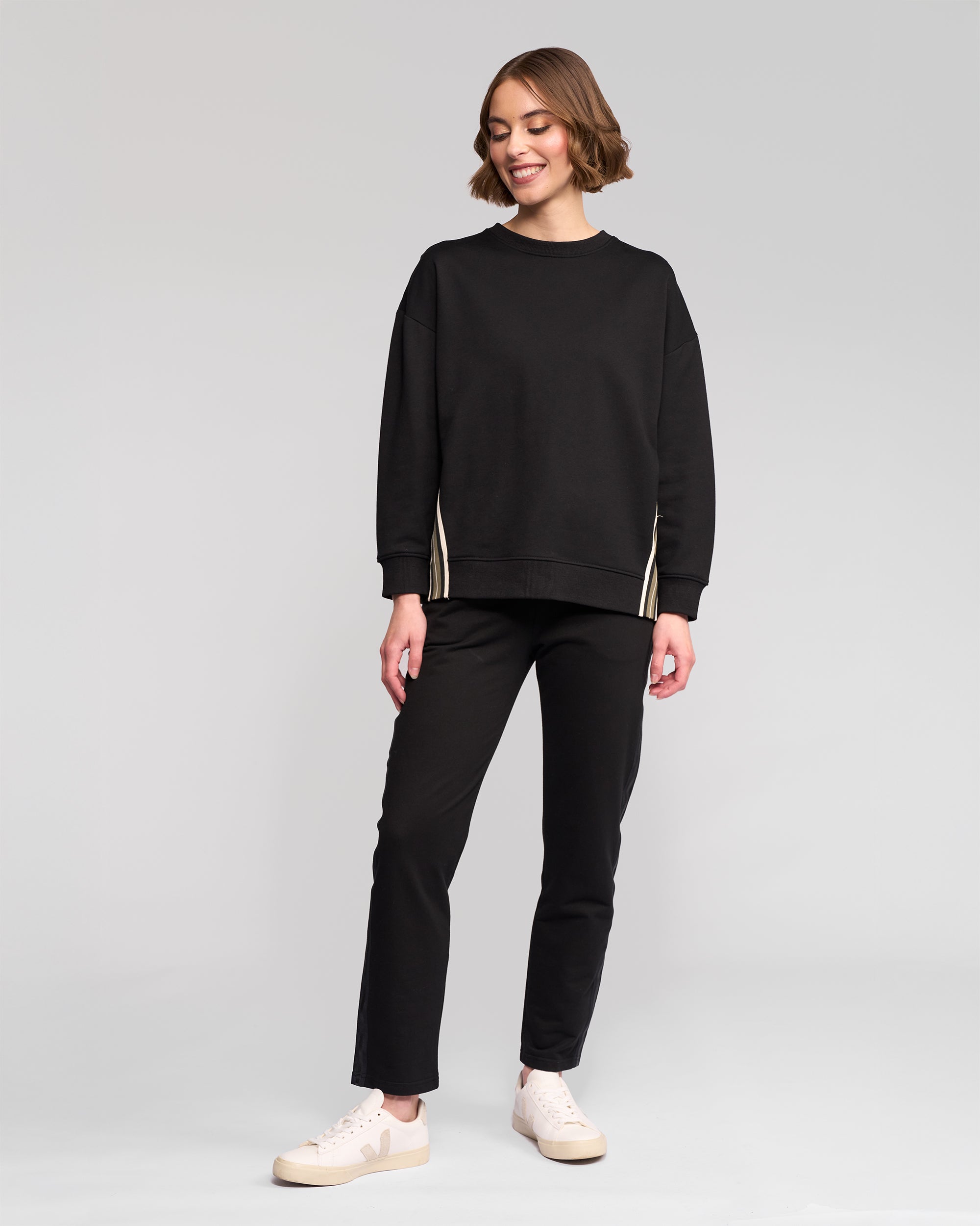 A woman with short brown hair smiles against a light gray background, wearing the Vassalli Zone Sweater in black, paired with black pants and light-colored sneakers.