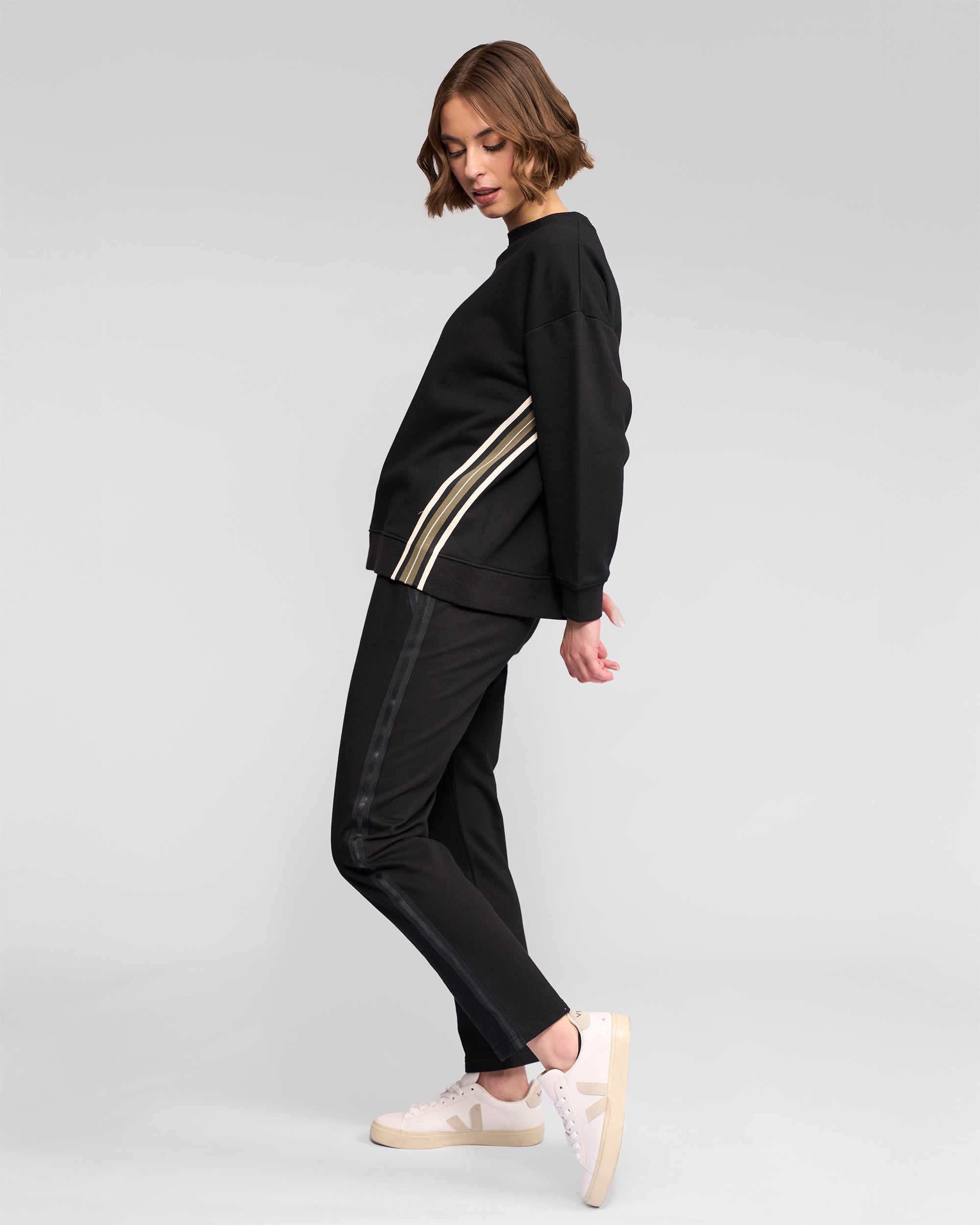 A woman poses sideways in a sporty Vassalli Zone Sweater - Black with white side stripes and matching black pants, paired with white sneakers against a plain light gray background.