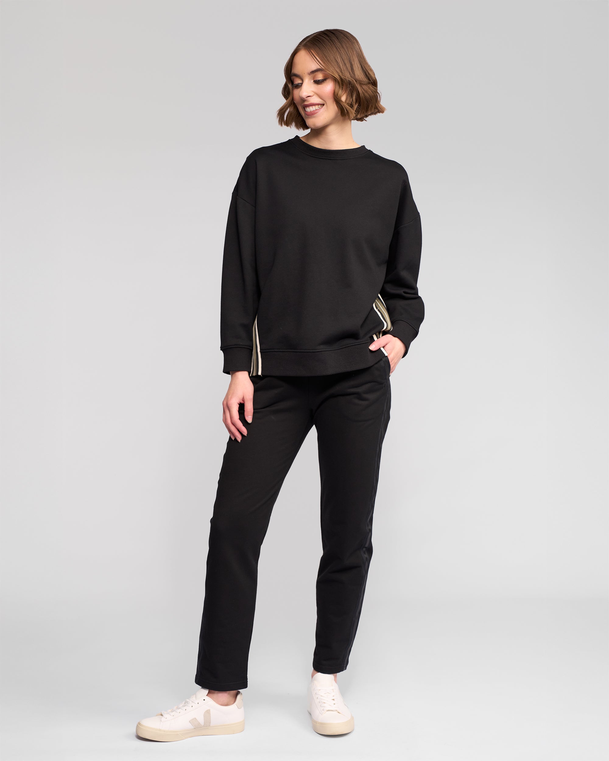 A person stands against a plain background, wearing the Vassalli Zone Sweater in black, paired with black pants and white sneakers with beige soles. They smile slightly, looking to the side with one hand in their pocket.