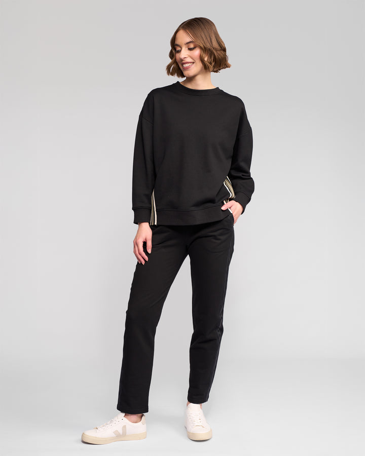 A person stands against a plain background, wearing the Vassalli Zone Sweater in black, paired with black pants and white sneakers with beige soles. They smile slightly, looking to the side with one hand in their pocket.