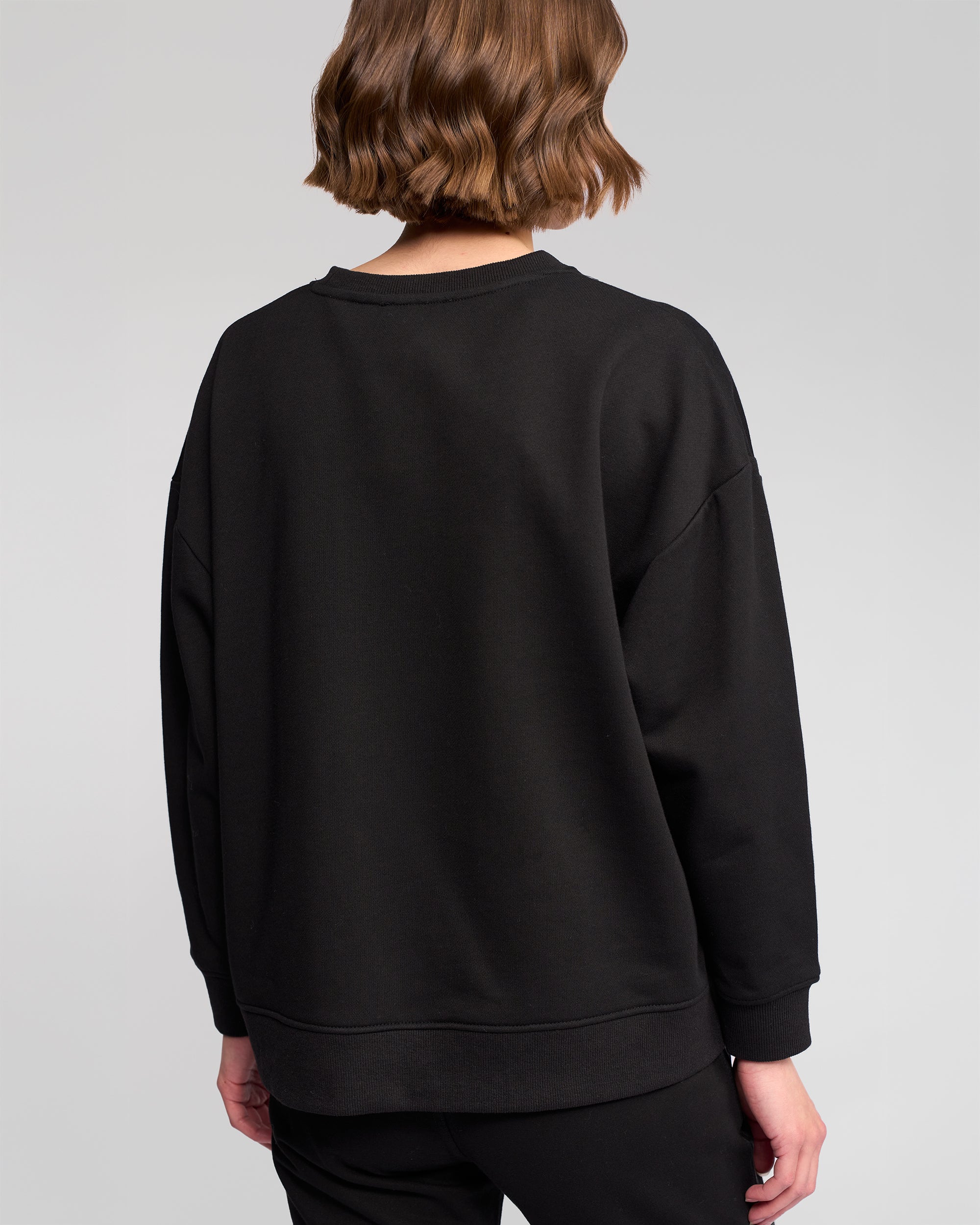A person with short, wavy brown hair is seen from behind, wearing the Vassalli Zone Sweater in black with matching black pants against a plain light gray background.