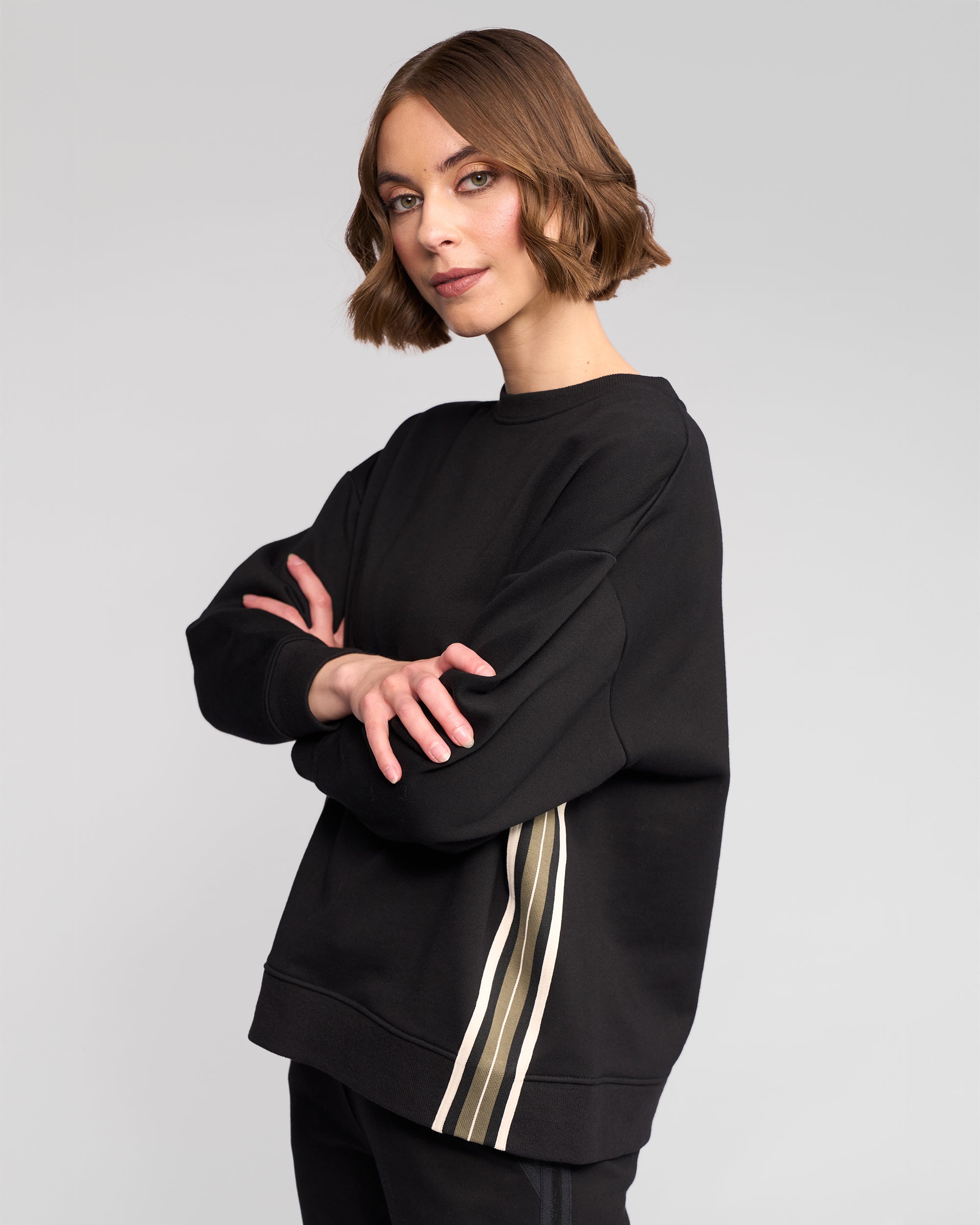 A woman with short brown hair stands confidently, arms crossed, wearing the Vassalli Zone Sweater in black with beige and white sleeve stripes, set against a plain light gray background.