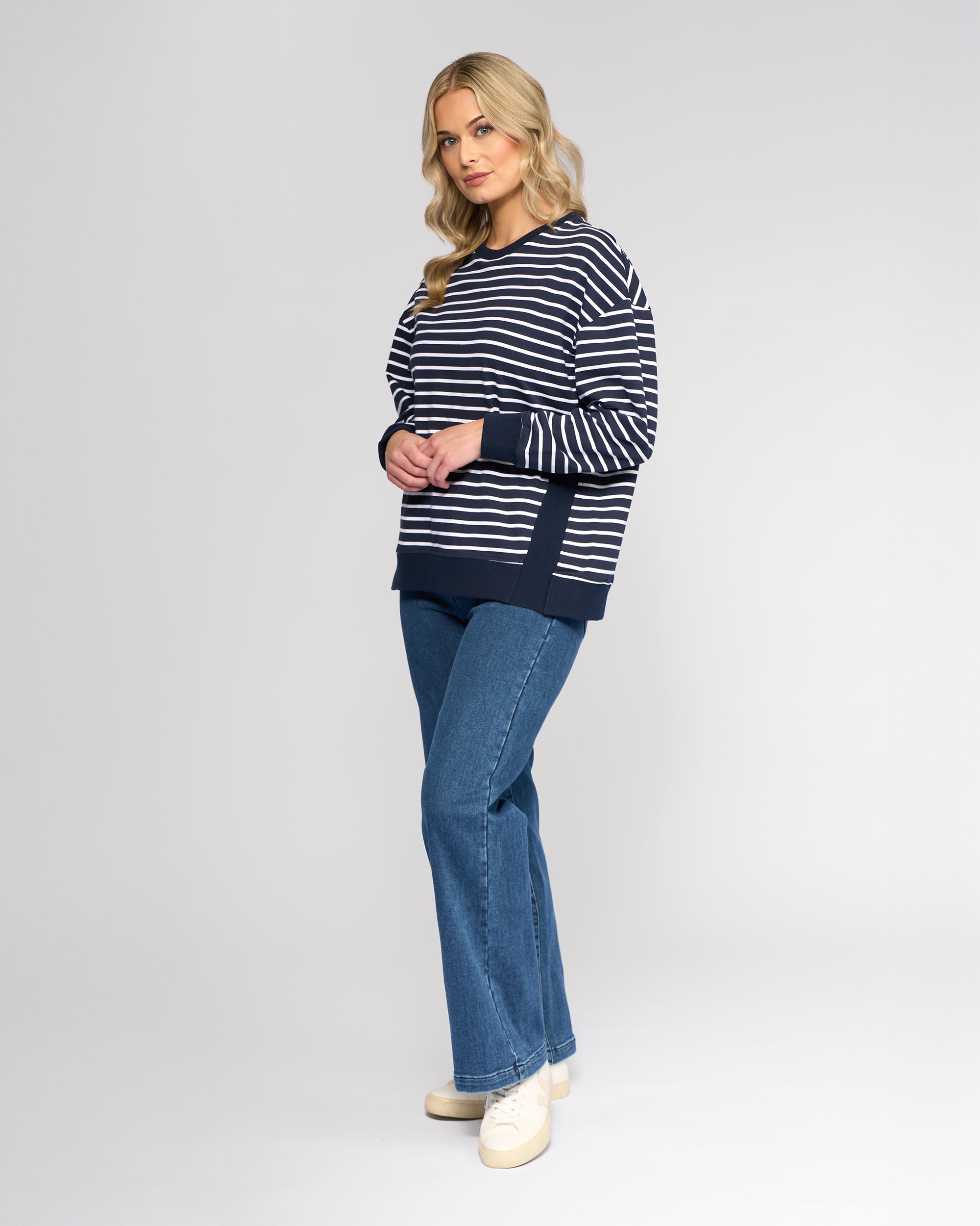 A woman with wavy blonde hair wears the Vassalli Zone Sweater in Ink Stripe, paired with blue jeans and cream shoes, standing against a plain light gray background.