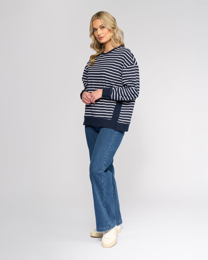 A woman with wavy blonde hair wears the Vassalli Zone Sweater in Ink Stripe, paired with blue jeans and cream shoes, standing against a plain light gray background.
