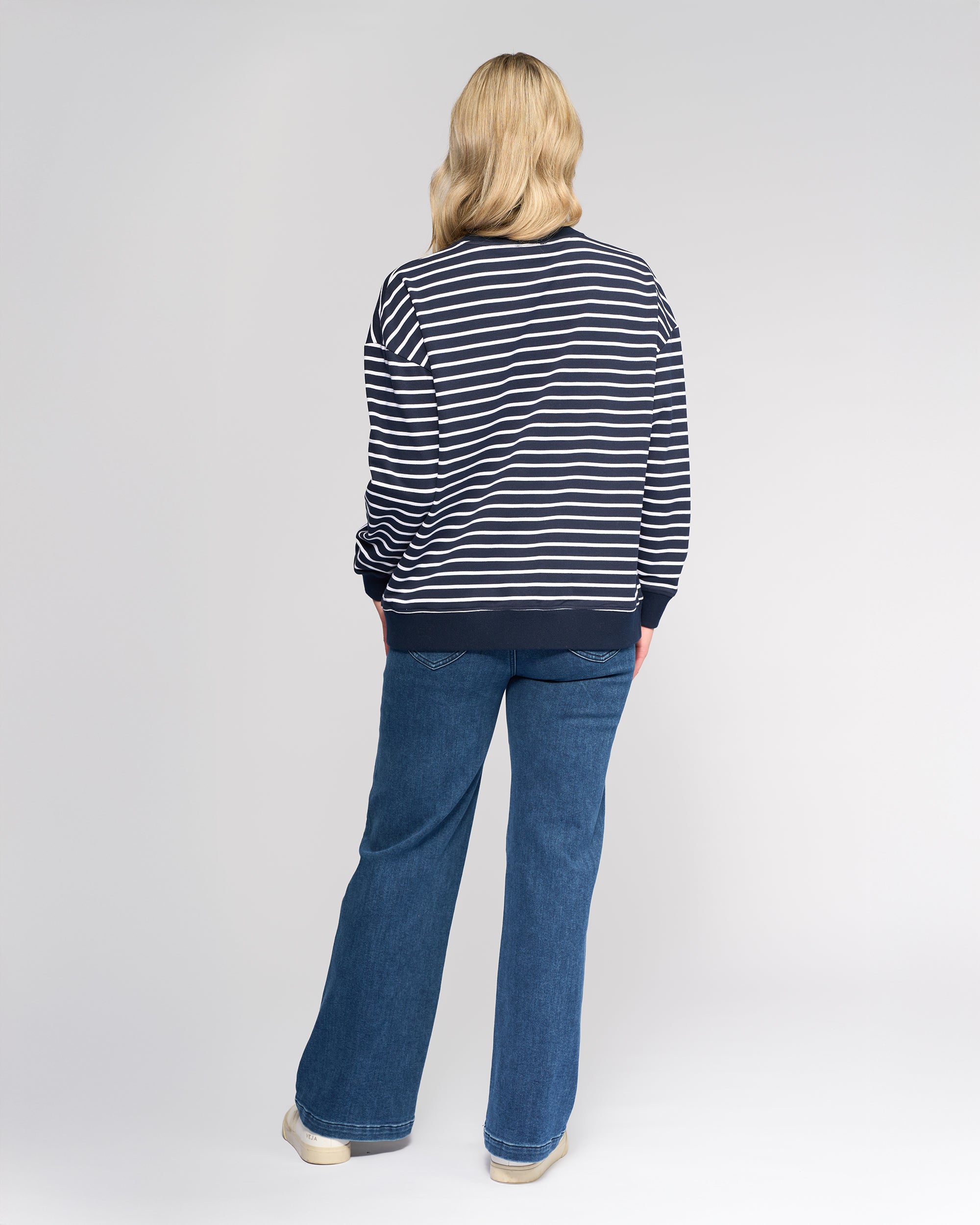 A person with blonde hair, seen from behind, wears the Vassalli Zone Sweater - Ink Stripe with blue jeans and white shoes, standing against a plain light background.