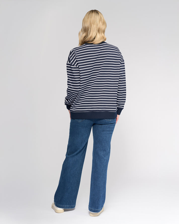 A person with blonde hair, seen from behind, wears the Vassalli Zone Sweater - Ink Stripe with blue jeans and white shoes, standing against a plain light background.