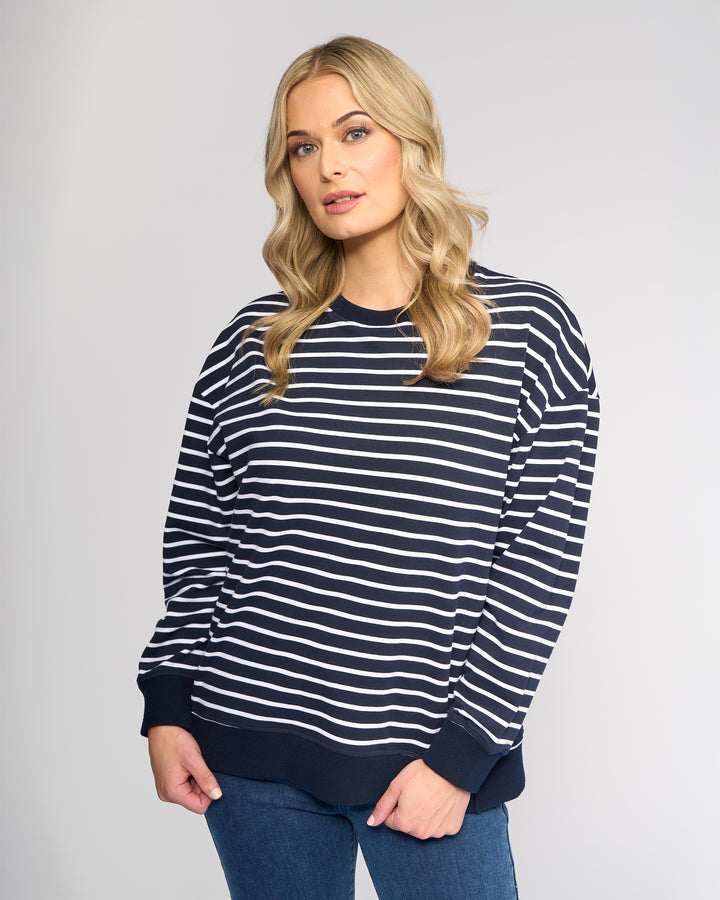 A woman with long blonde hair wears the Vassalli Zone Sweater - Ink Stripe with blue jeans, standing against a plain light gray background.