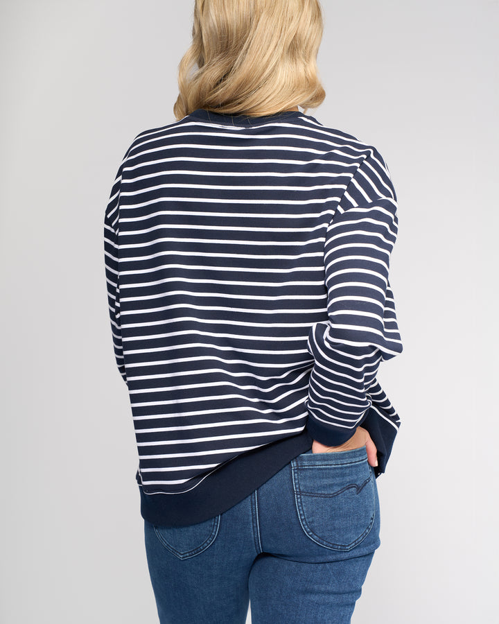 A person with blonde hair is seen from behind wearing the Vassalli Zone Sweater - Ink Stripe, a relaxed-fit navy and white cotton knit crew neck, paired with blue jeans, against a plain light background.