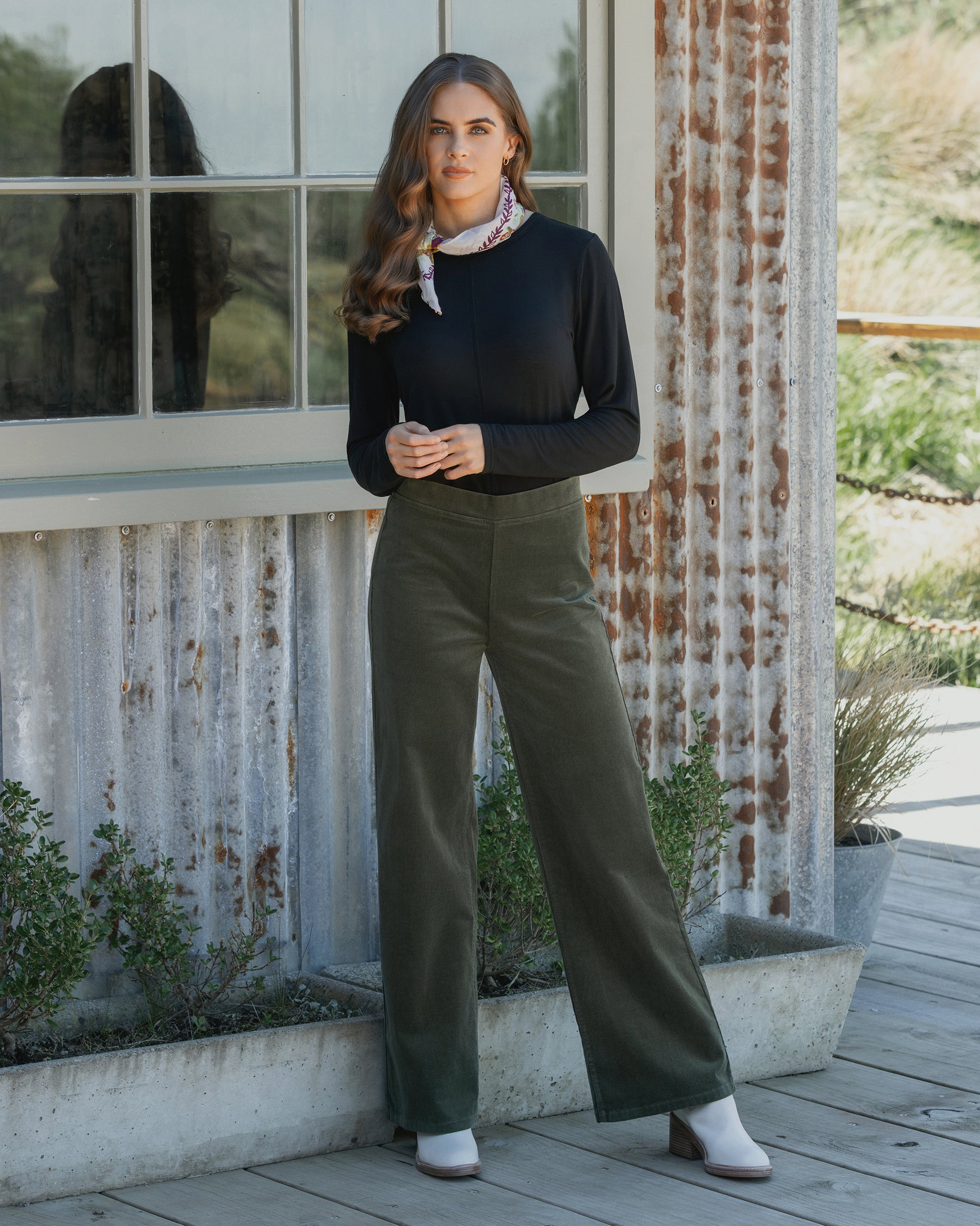 A woman with long brown hair stands before a rustic metal building, wearing a black top, white boots, patterned scarf, and Vassalli’s Loft Cord Pull On wide-leg pants in Deep Sage. Shrubs grow at her feet.
