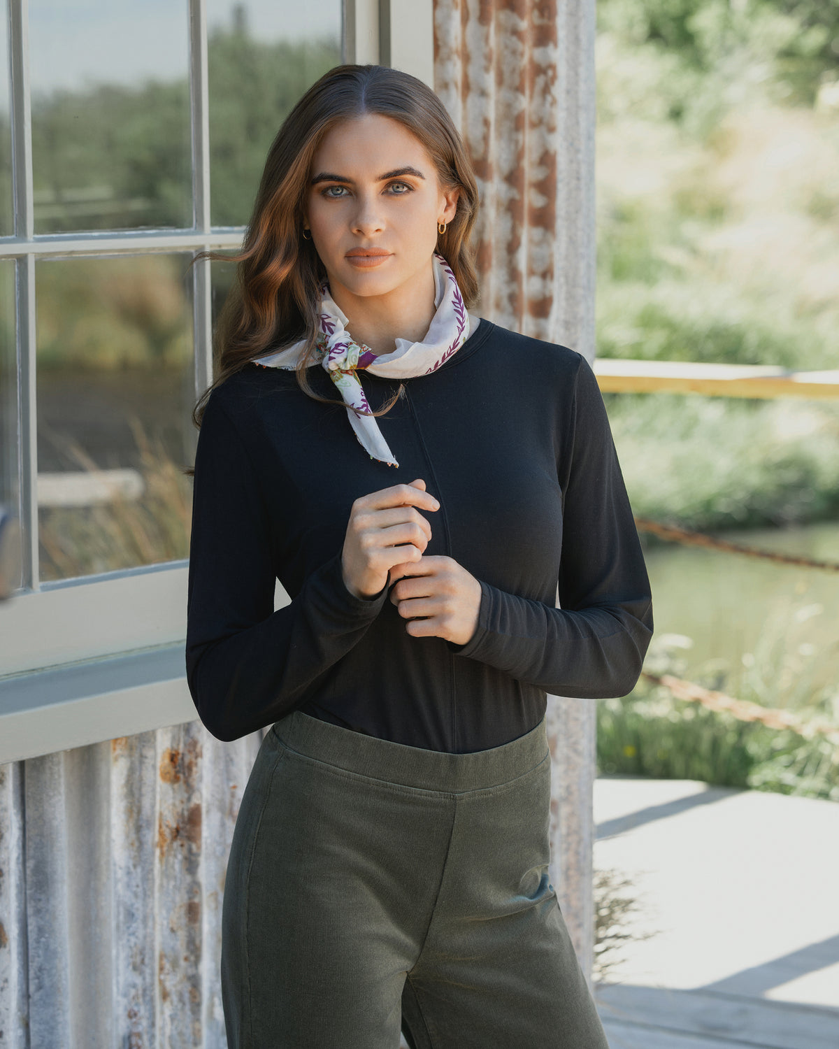 A woman with long brown hair stands outside by a corrugated metal wall and window, wearing the Vassalli Layer Top - Black. She pairs it with olive pants and a floral scarf, looking directly at the camera with a neutral expression.