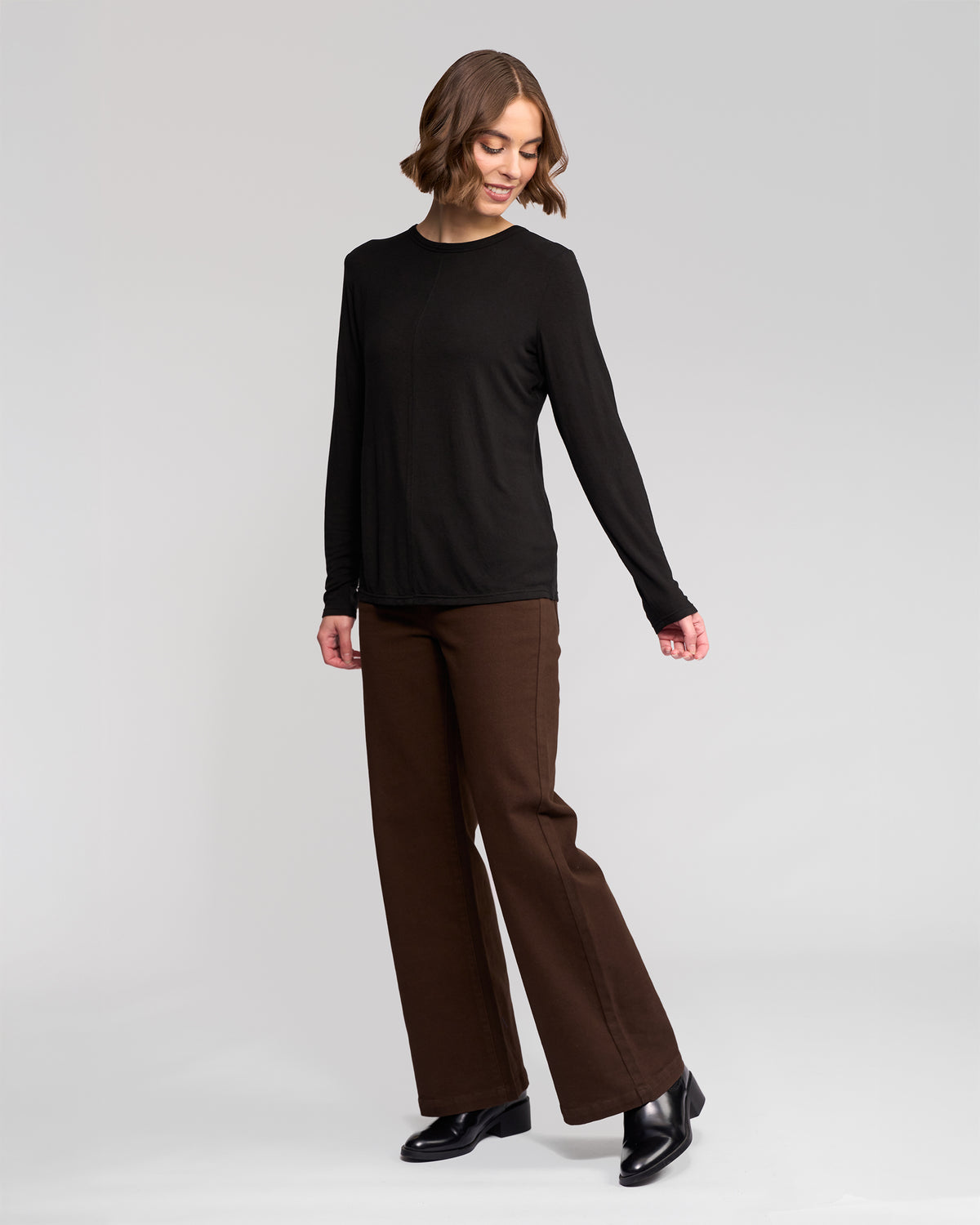 A woman with short brown hair wears the Vassalli Layer Top in black, paired with brown wide-leg pants and black ankle boots. She stands on a plain light background, looking down and smiling.