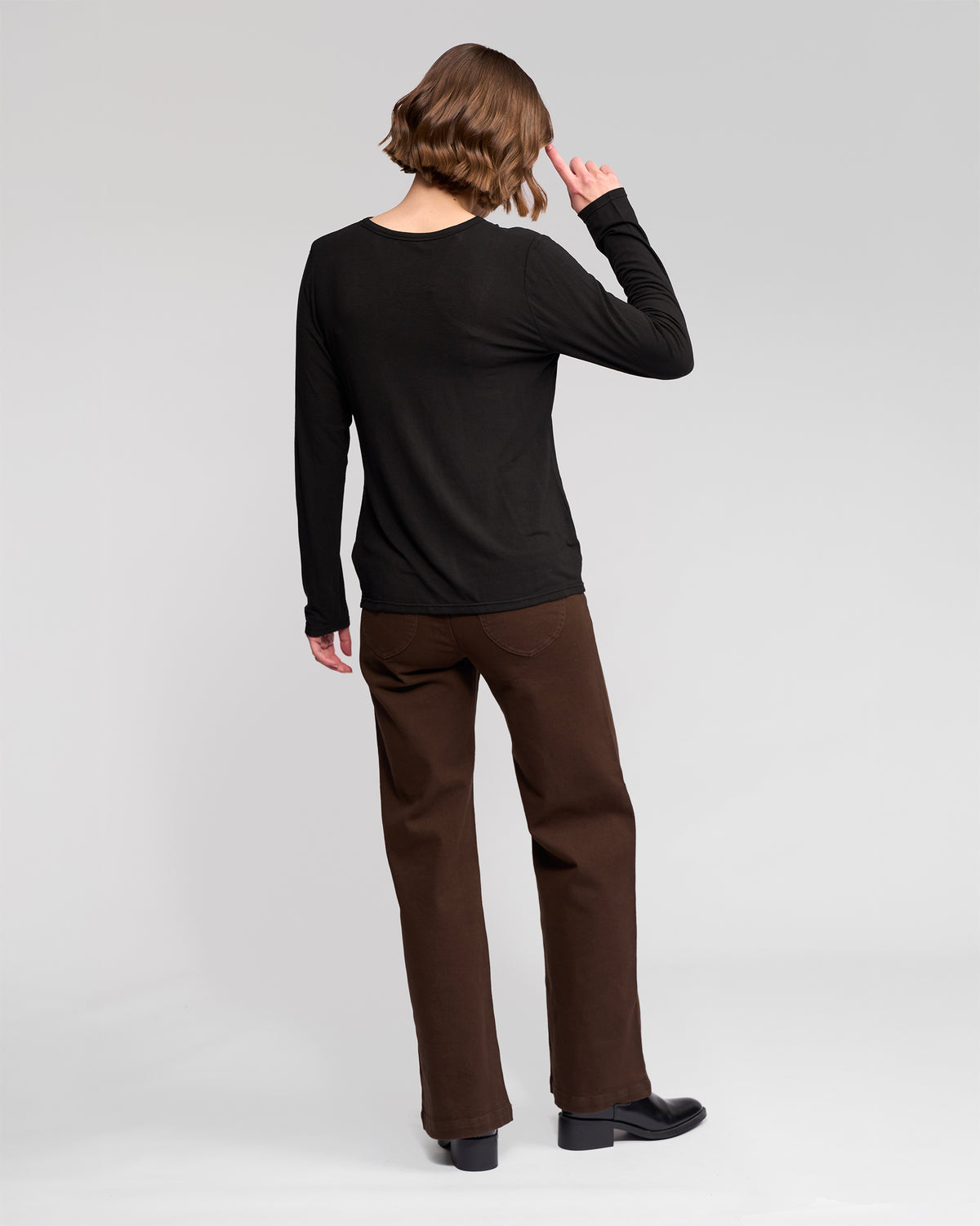 A person with short, wavy hair faces away, wearing the Vassalli Layer Top - Black paired with brown pants and black shoes against a plain light background.
