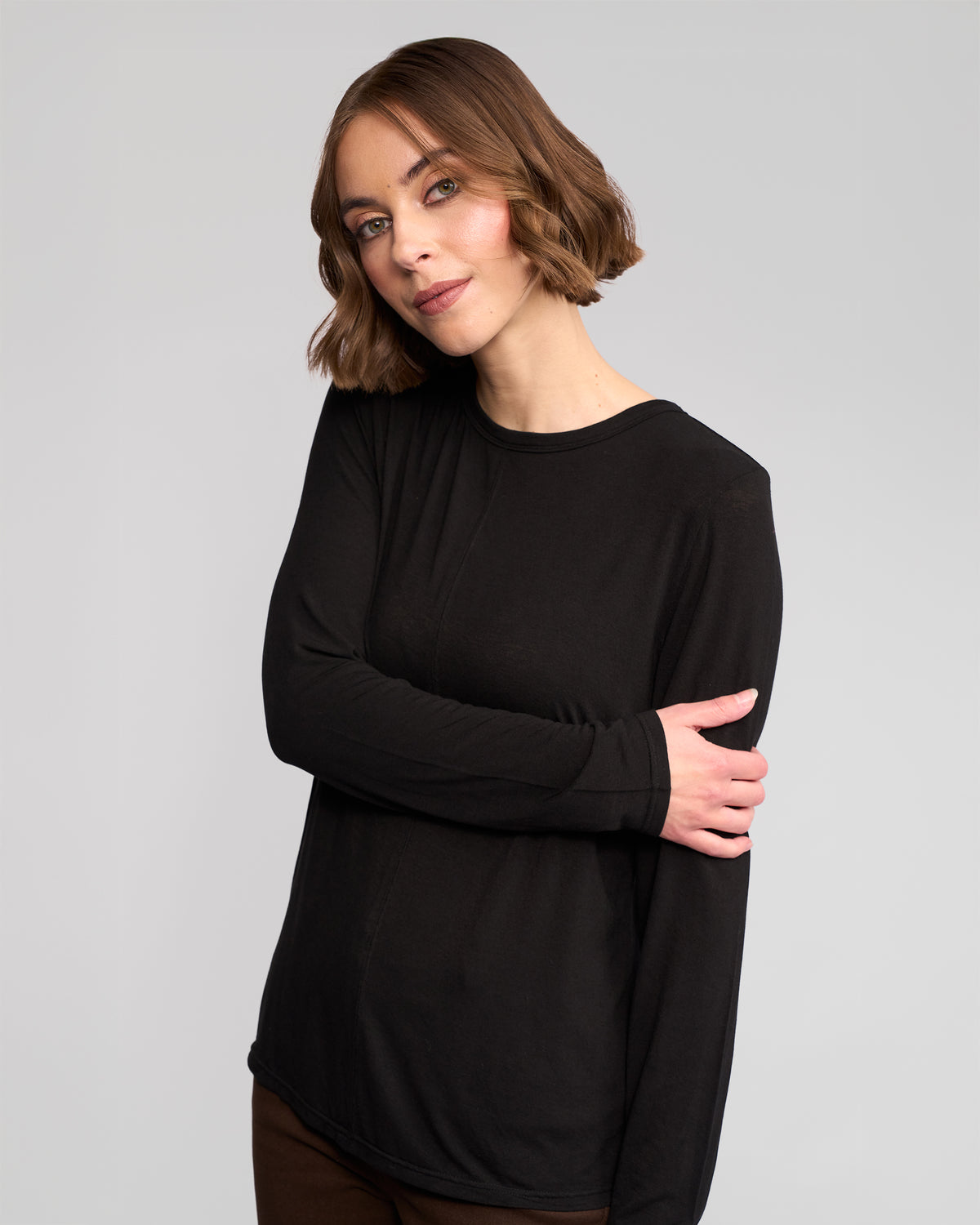 A woman with short, wavy brown hair stands against a light gray background, wearing the Vassalli Layer Top - Black and brown pants. With one arm crossed and a gentle expression, she showcases this timeless wardrobe essential.