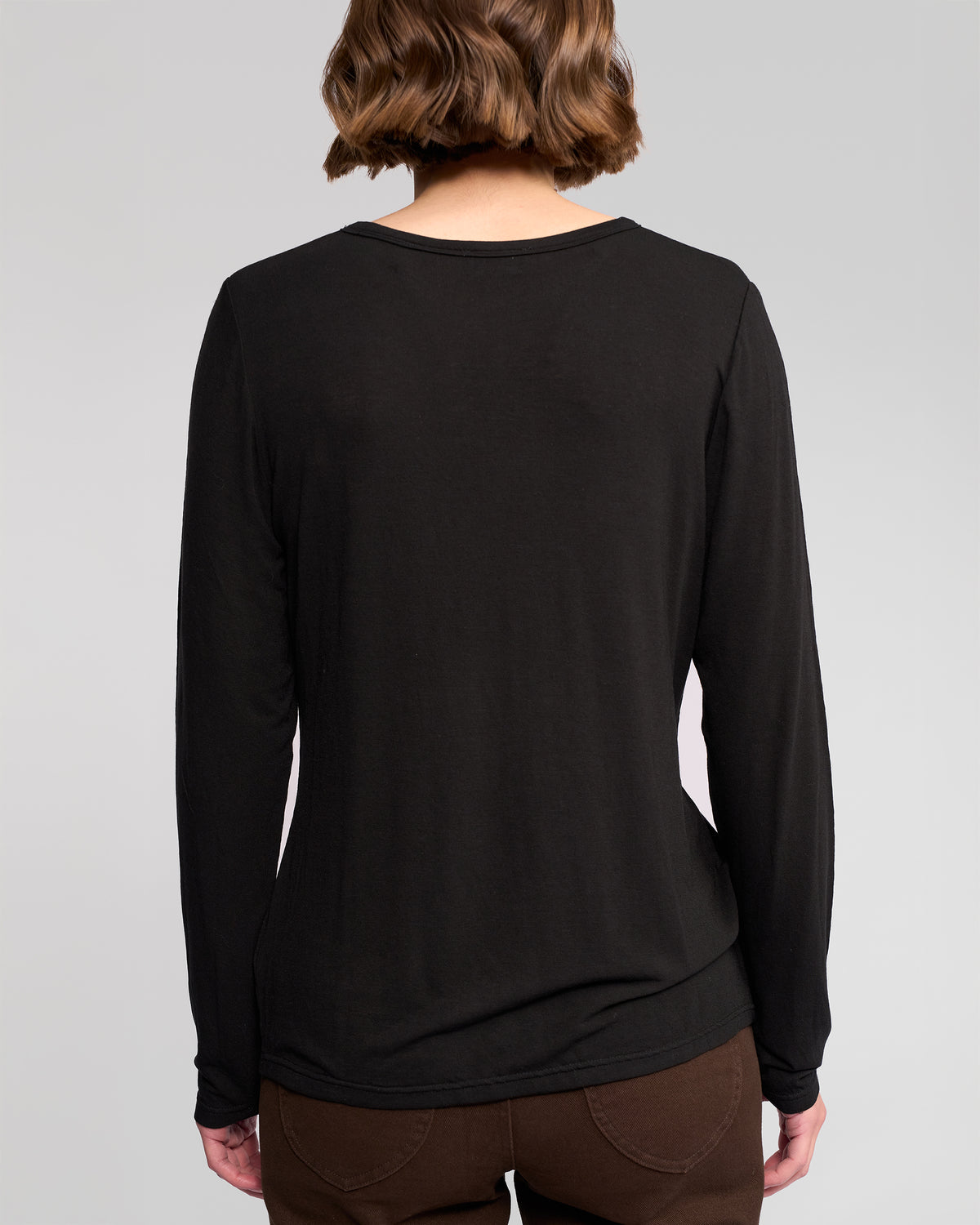 A person with short, wavy brown hair is seen from behind wearing the Vassalli Layer Top in black with brown pants, against a plain light background.