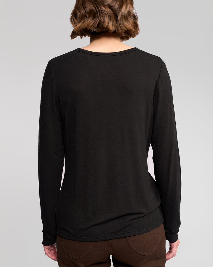 A person with short, wavy brown hair is seen from behind wearing the Vassalli Layer Top in black with brown pants, against a plain light background.