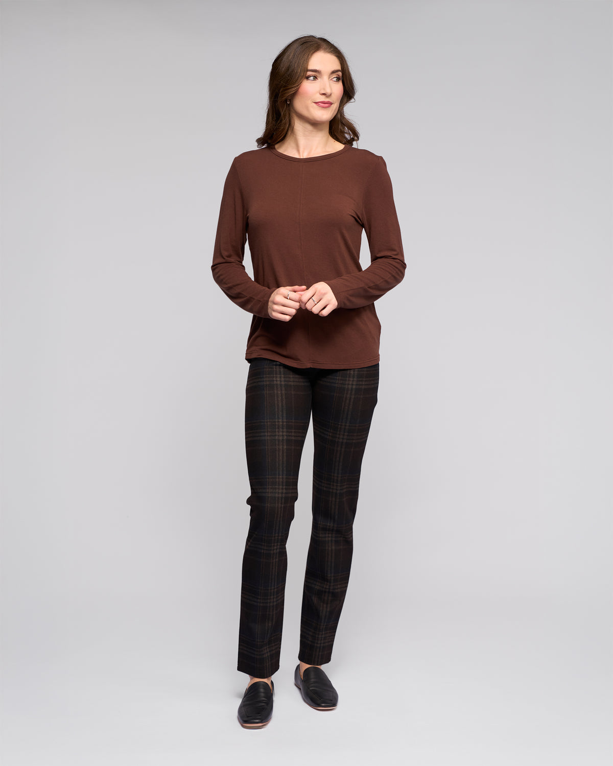 A woman stands in a studio against a plain background, wearing the Vassalli Layer Top in chocolate, dark plaid pants, and black slip-on shoes. She looks slightly to the side with a soft smile, hands loosely clasped.